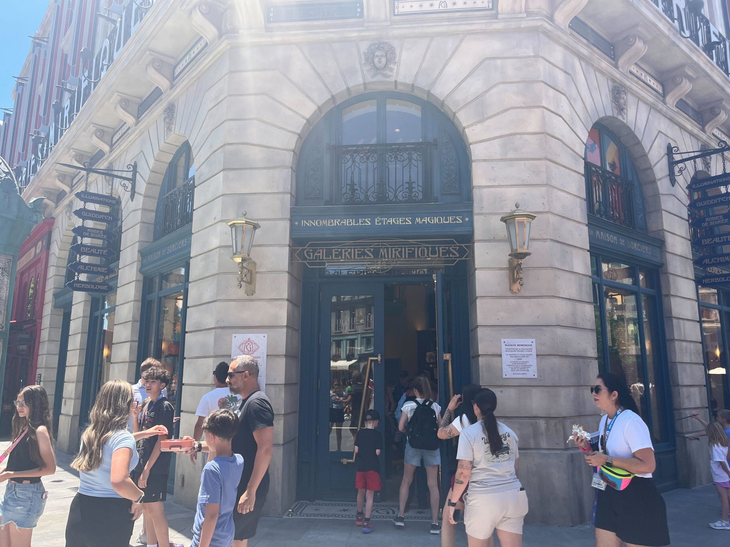 People stand and walk outside Les Galeries Mirifiques on a sunny day, with signs on the building and blue skies above.