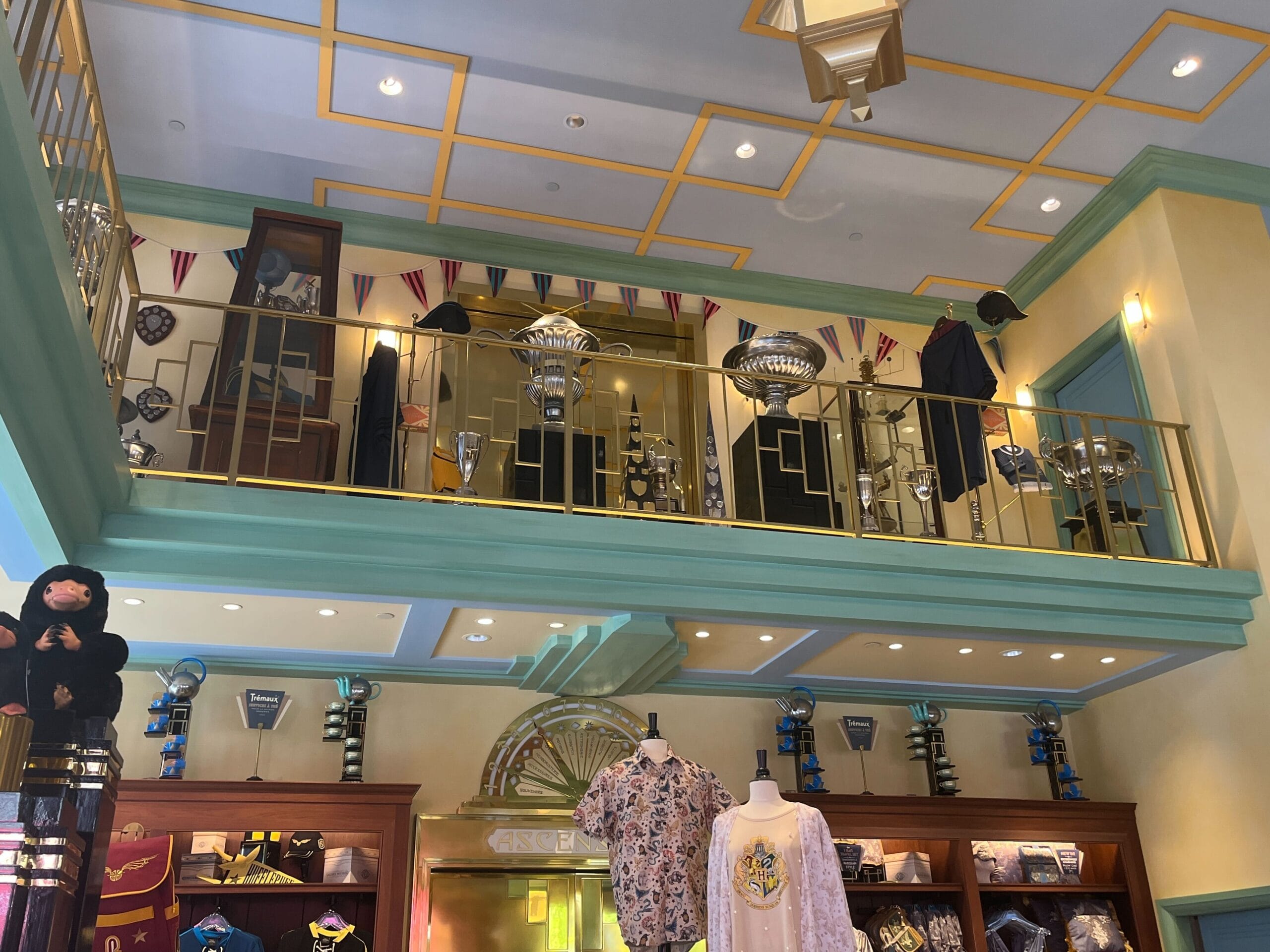 A shop interior at Les Galeries Mirifiques features school-themed decor, uniforms, trophies, and shirts under gold railings.