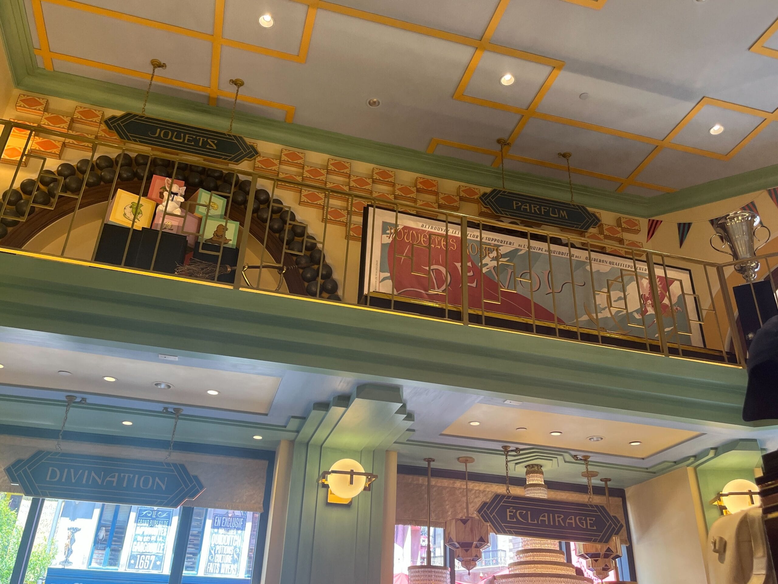 Interior of Les Galeries Mirifiques shop with green walls, labeled signs, and colorful boxes on display shelves near the ceiling.