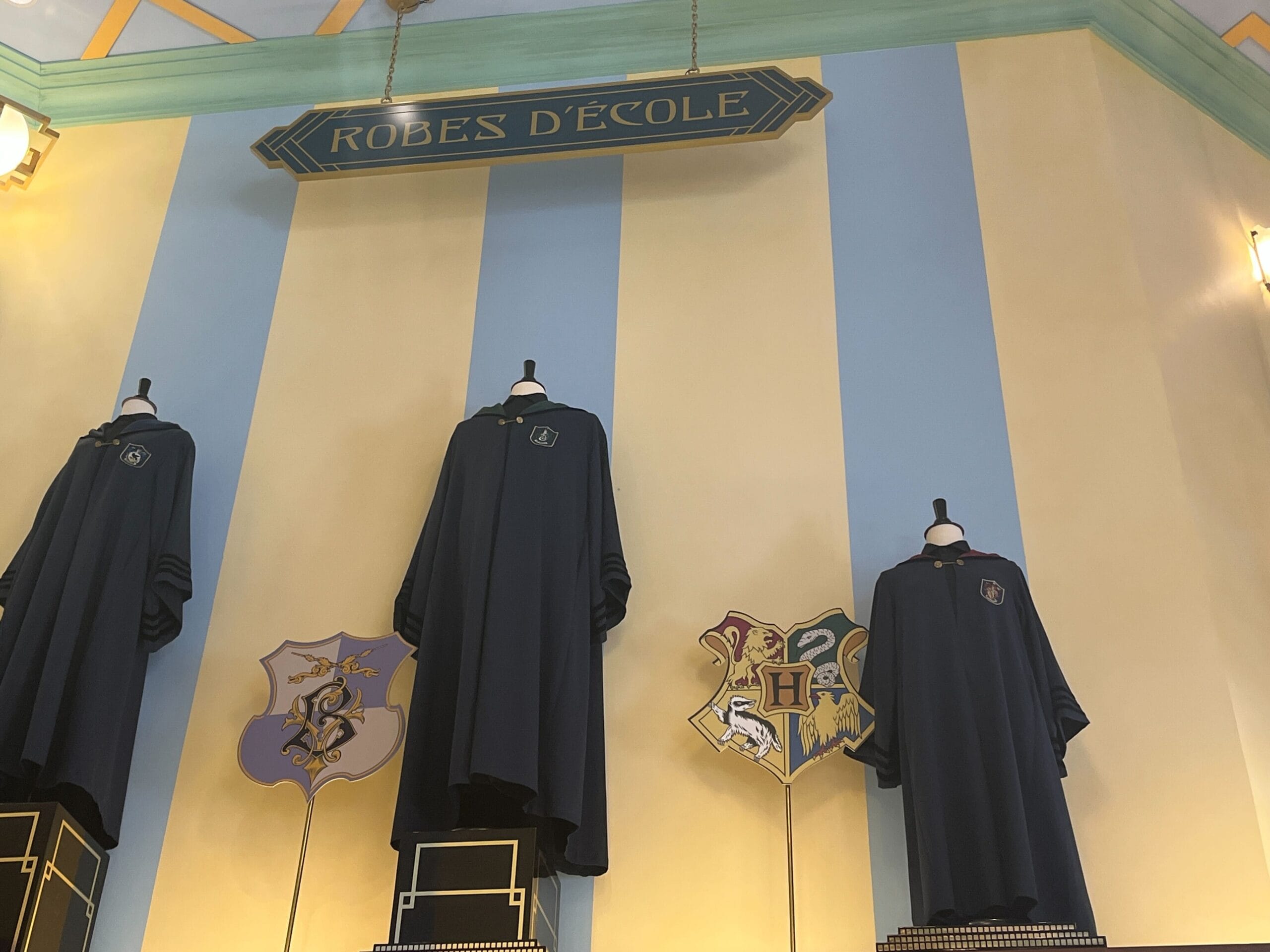 Three school robes on mannequins beneath Hogwarts house crests at Les Galeries Mirifiques, with a sign reading "Robes d'École.