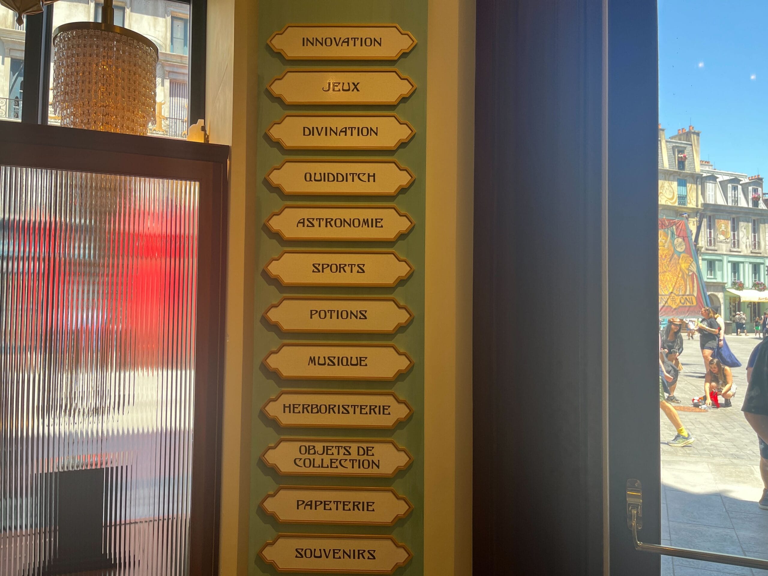 Vertical sign inside Les Galeries Mirifiques with French sections: "Sports," "Potions," and "Souvenirs.