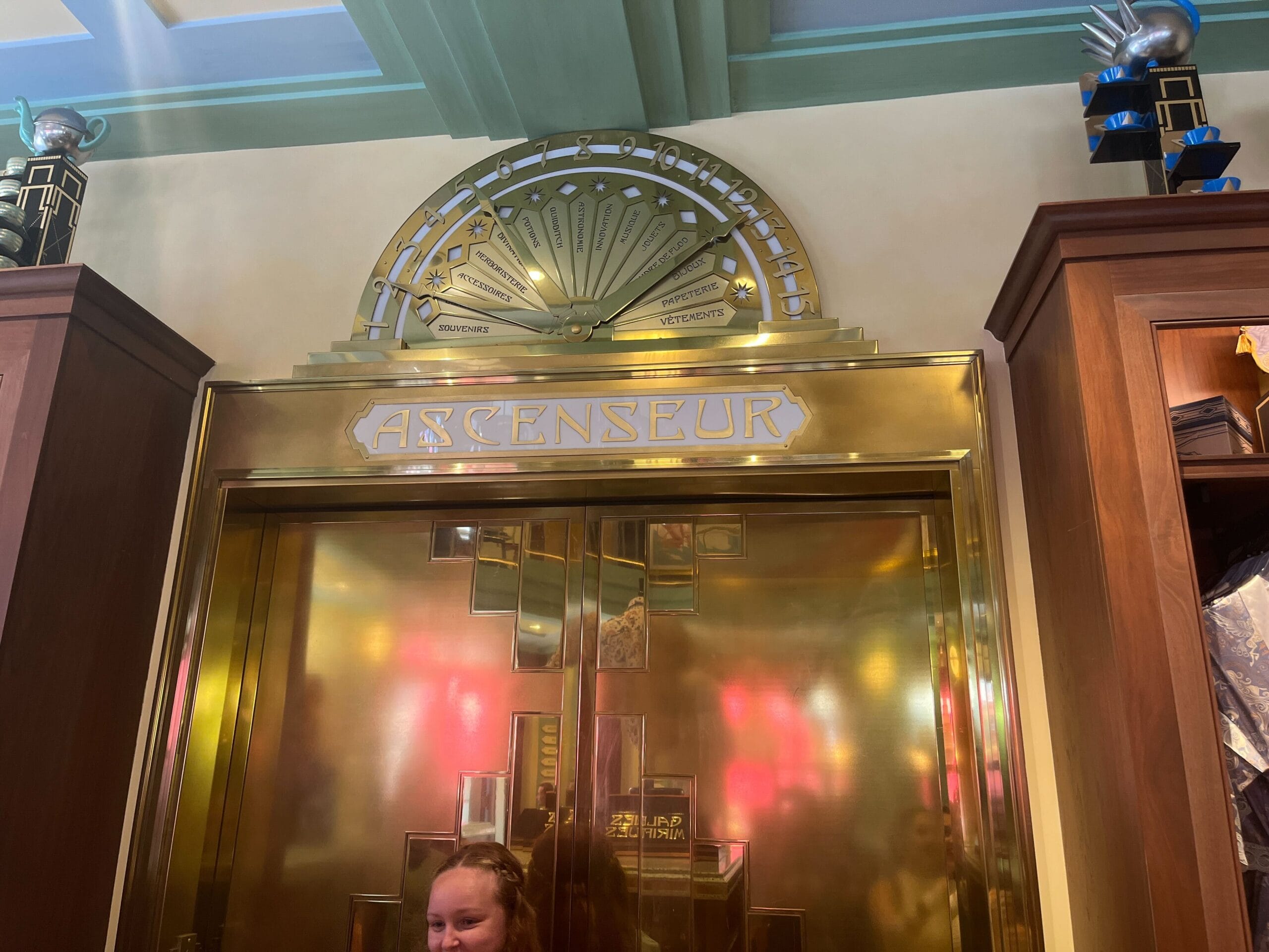 A golden elevator with Art Deco design at Les Galeries Mirifiques; "ASCENSEUR" gleams above as a person stands in front.