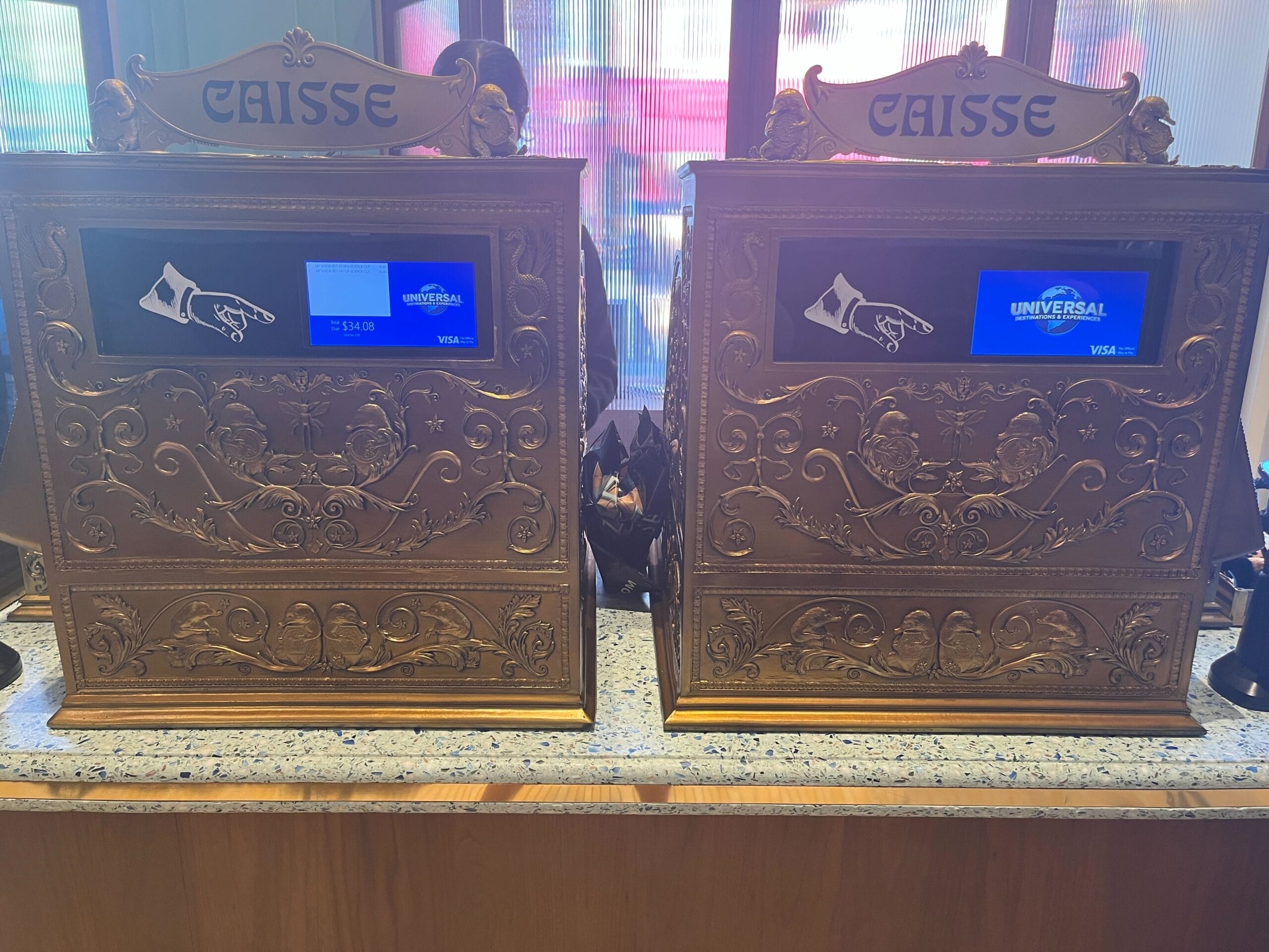 Two ornate, gold cash registers labeled "CAISSE" at Les Galeries Mirifiques, on a marble countertop.