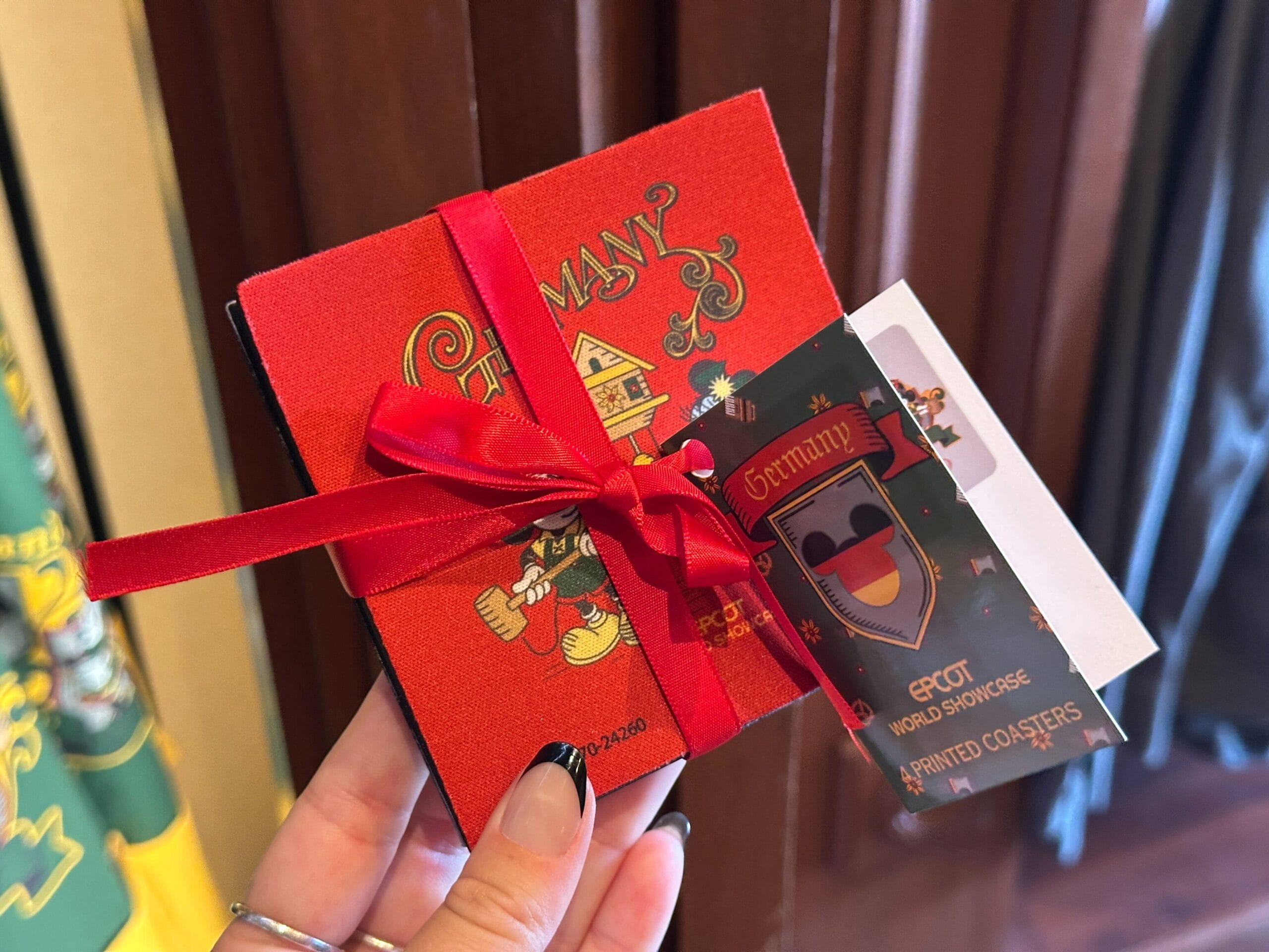 A hand holds Germany Pavilion merchandise: red coasters with Mickey Mouse and an EPCOT World Showcase tag, tied with a ribbon.