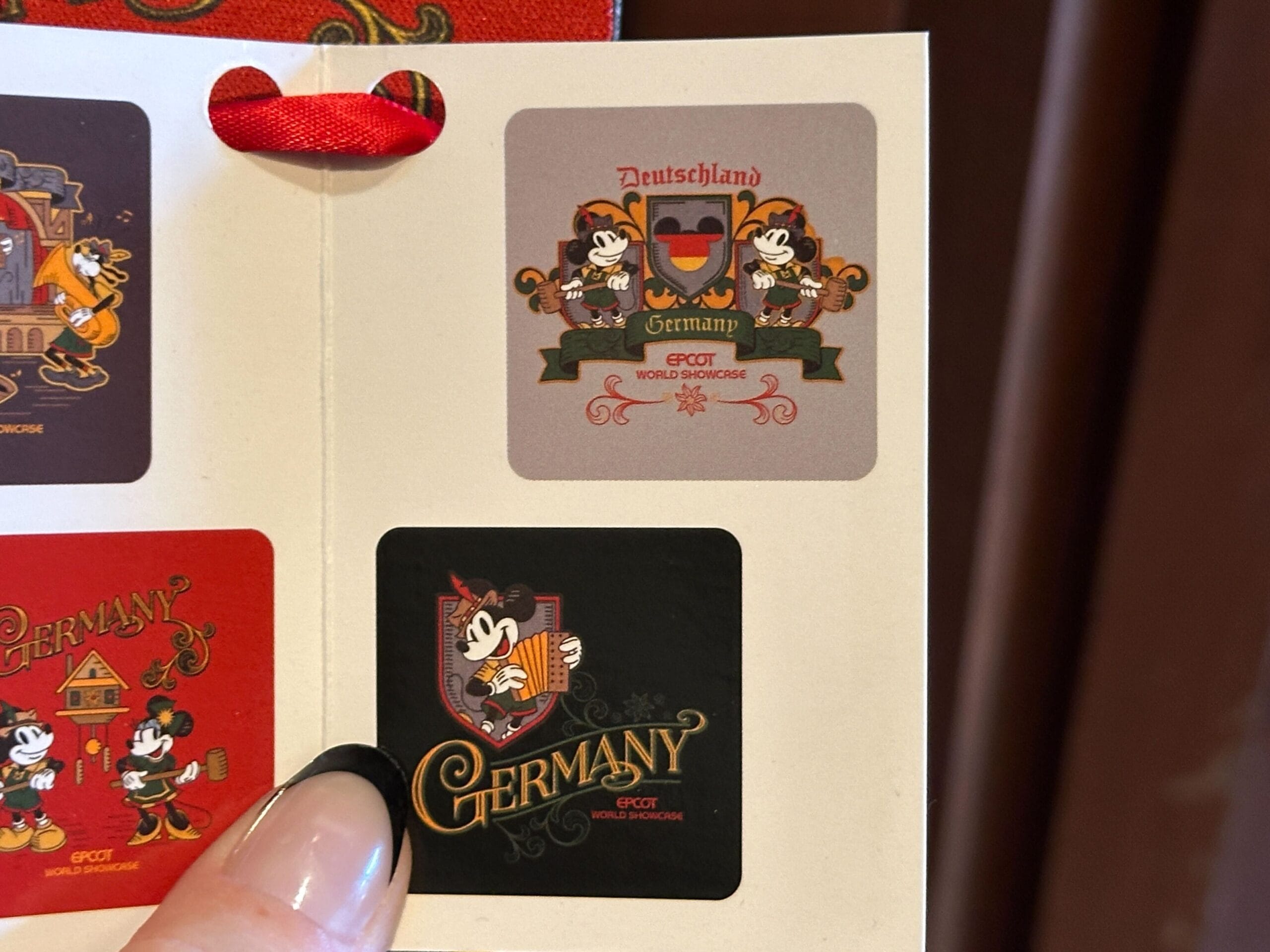A hand holds merchandise—a booklet with two Germany Pavilion-themed Mickey Mouse stickers from EPCOT World Showcase.