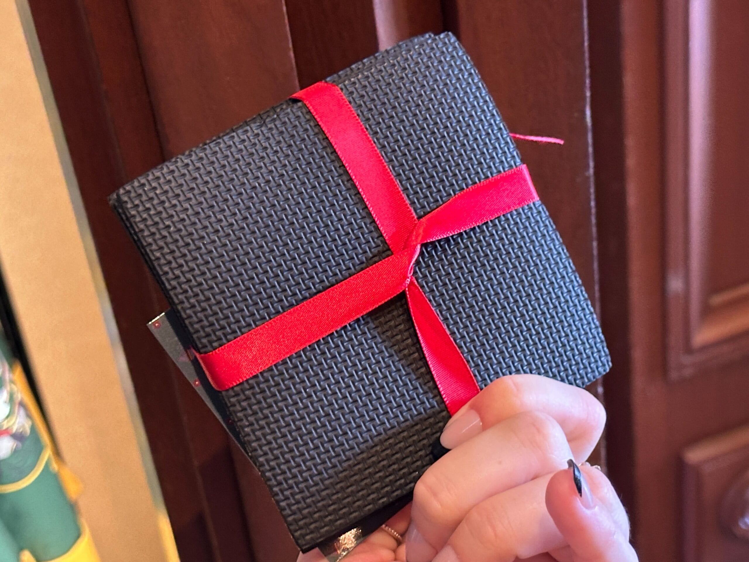A hand holds Germany Pavilion merchandise—a textured black wallet tied with a red ribbon—before a wooden background.