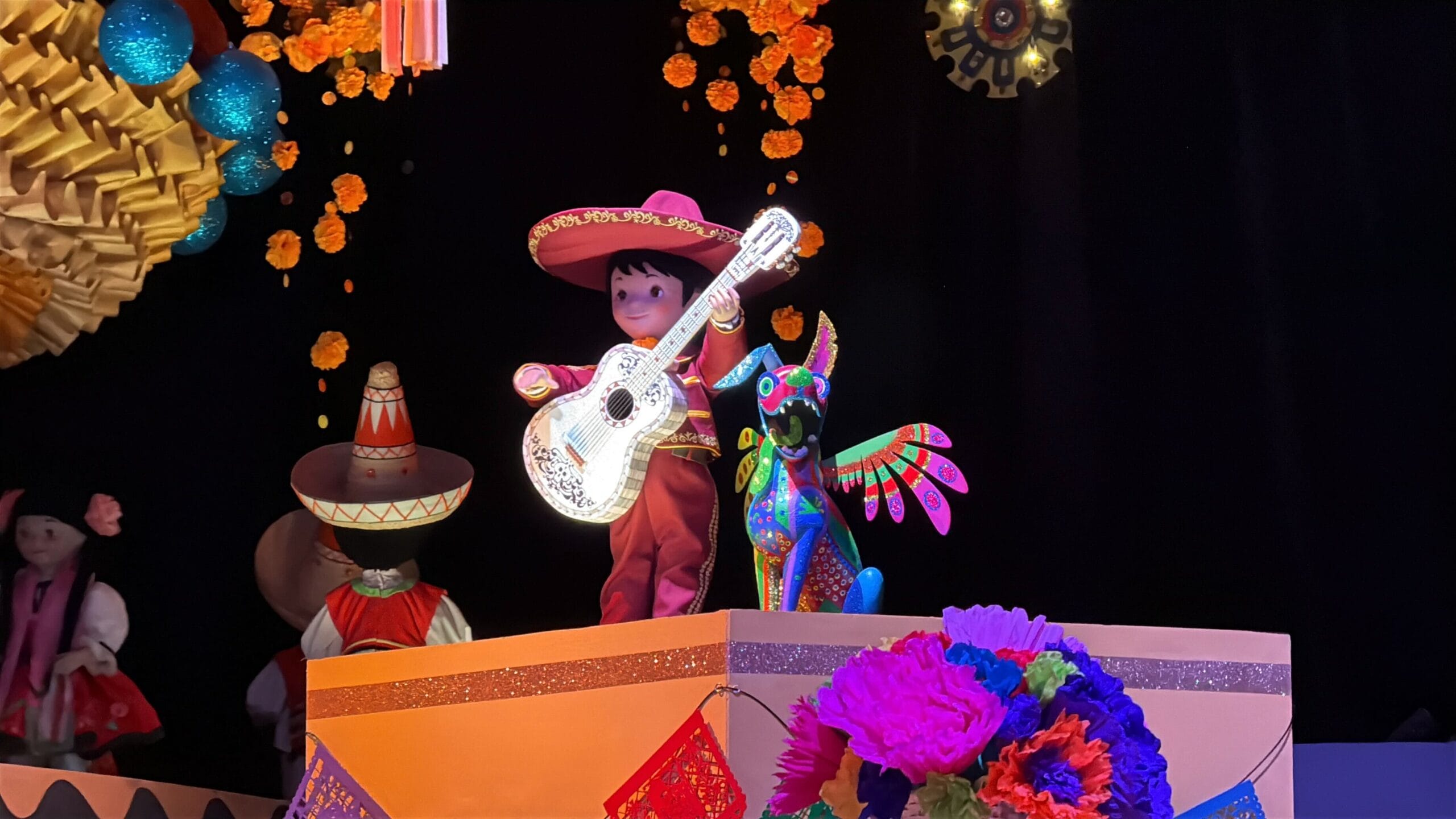 A boy in a sombrero plays guitar beside Coco figures on a festive stage, inspired by Disneyland 70th celebrations.