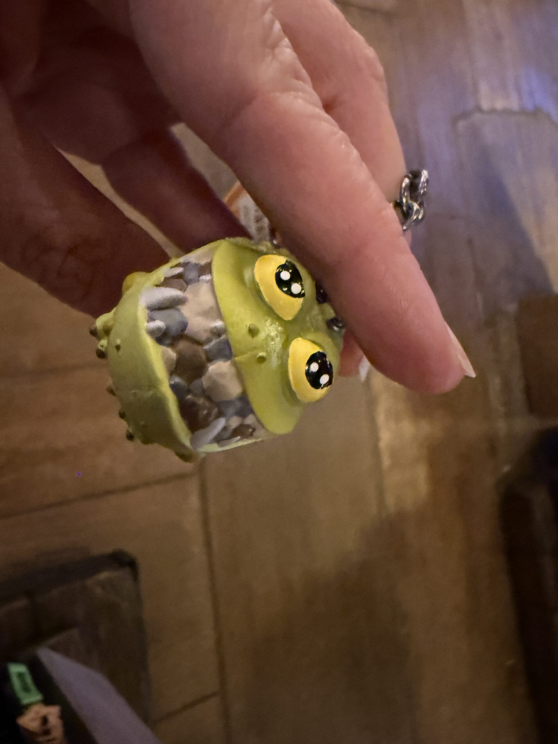 A hand holds a small, green, cartoonish creature keychain from Hiccup's Work Shop with big eyes and a toothy grin.