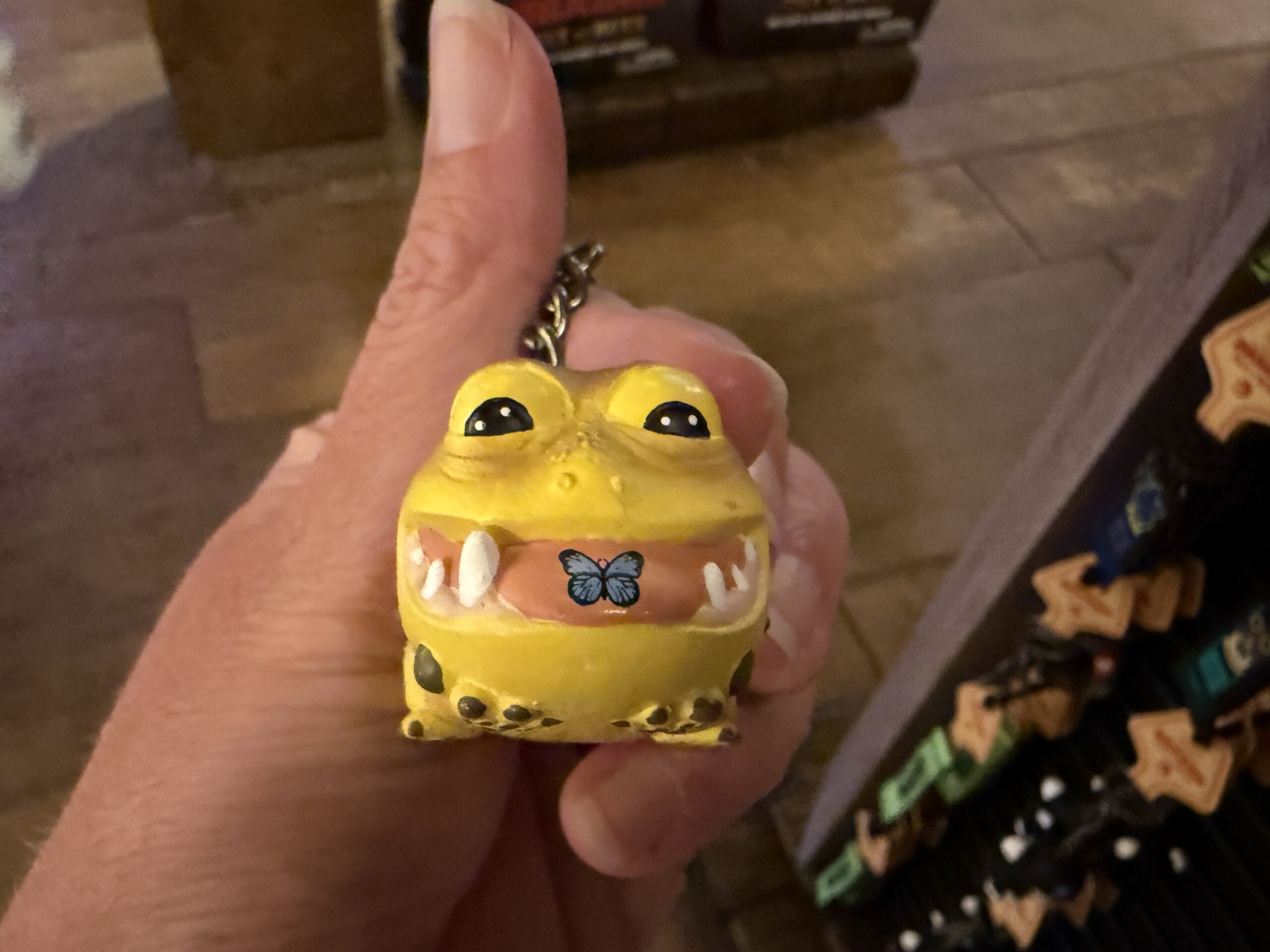 A hand holds a yellow frog keychain from Hiccup's Work Shop, with big eyes, teeth, and a blue butterfly on its mouth.