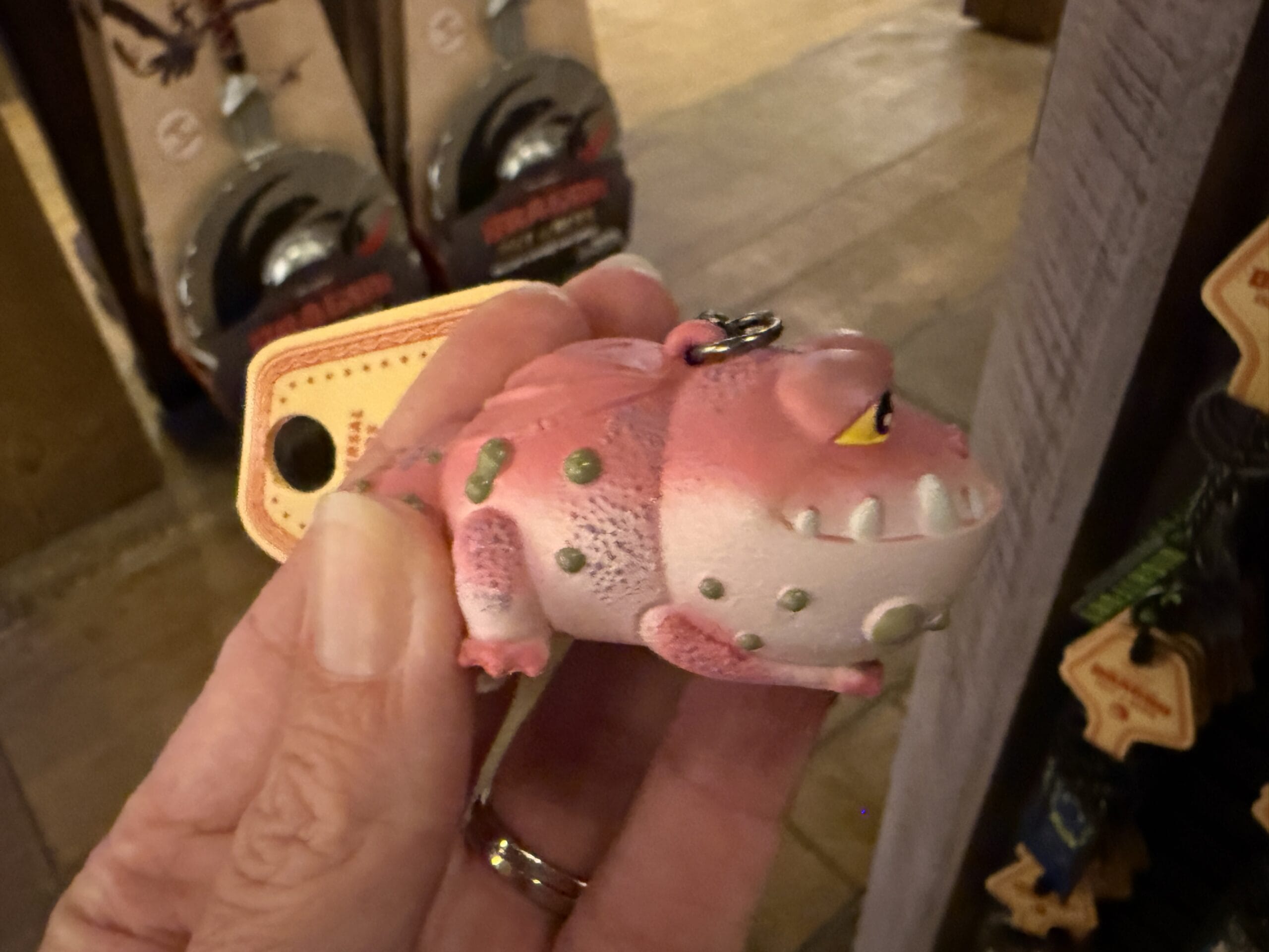 A hand holds a small, pink dragon-like keychain toy from Hiccup's Work Shop in a store.