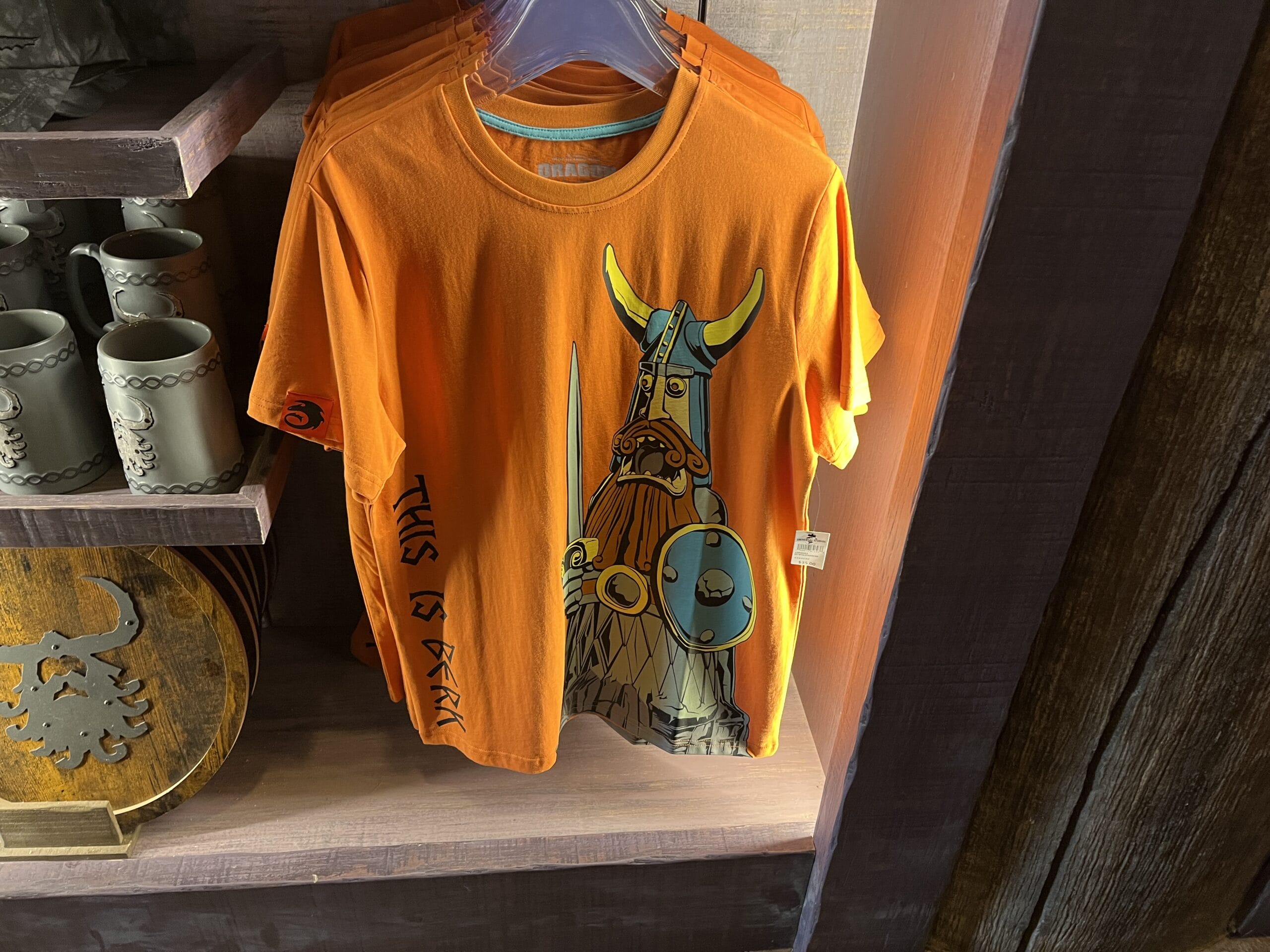 Orange t-shirt featuring a cartoon Viking warrior from Hiccup's Work Shop, displayed on a wooden shelf.