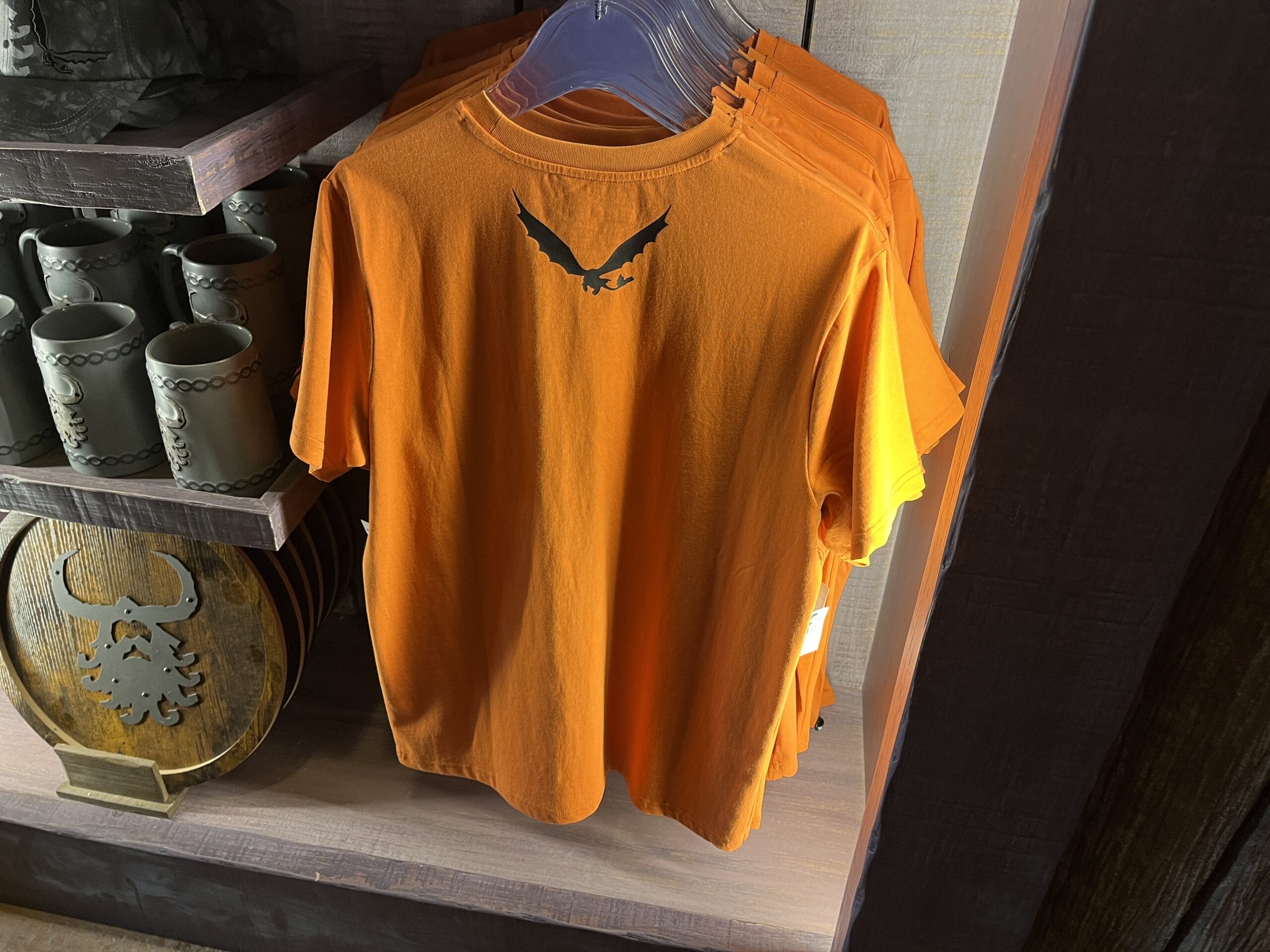 Bright orange t-shirt from Hiccup's Work Shop, featuring a black silhouette of a flying dragon on a store rack.