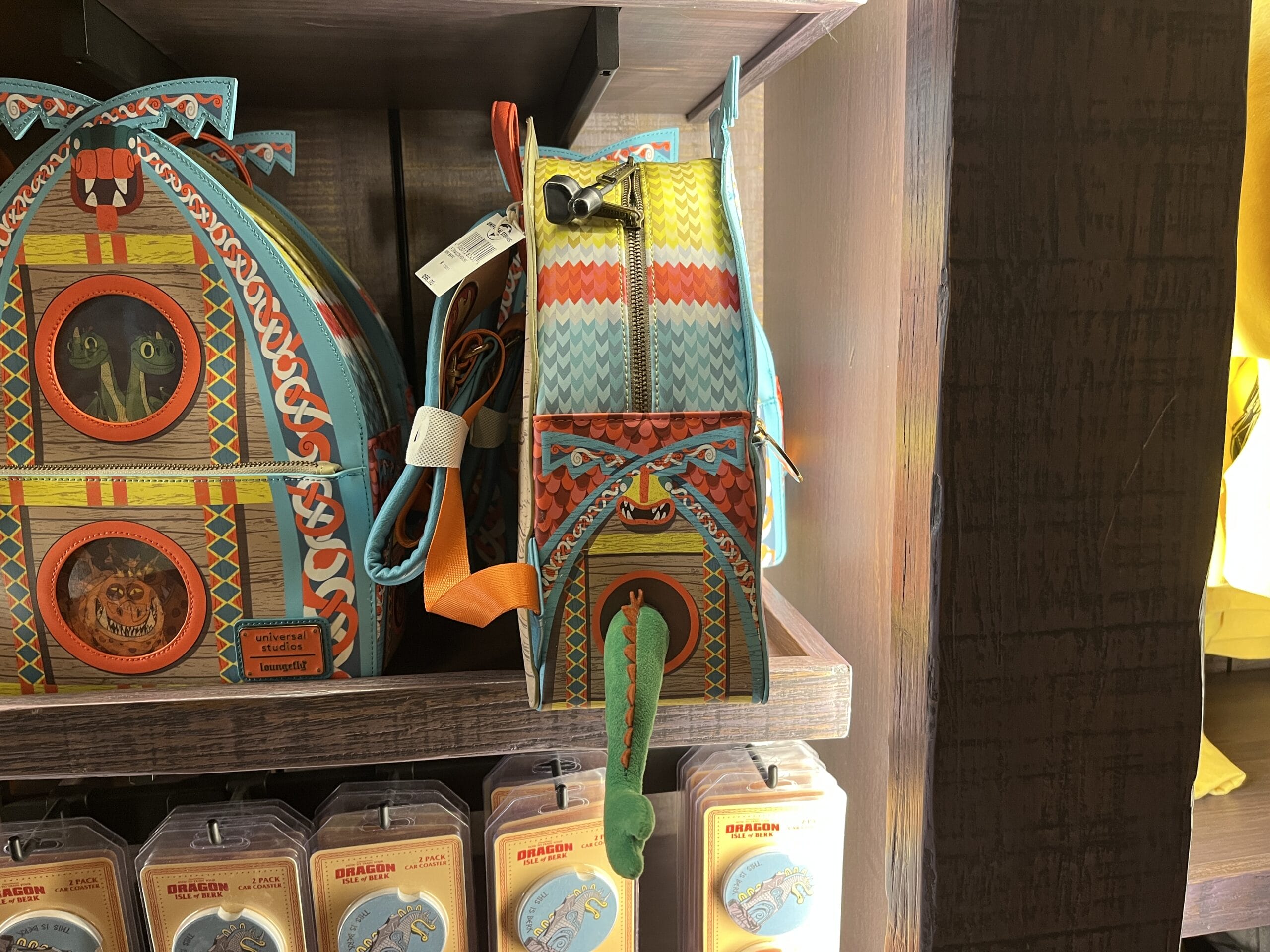 Colorful dragon-themed backpacks from Hiccup's Work Shop are displayed on wooden shelves, with keychains below.