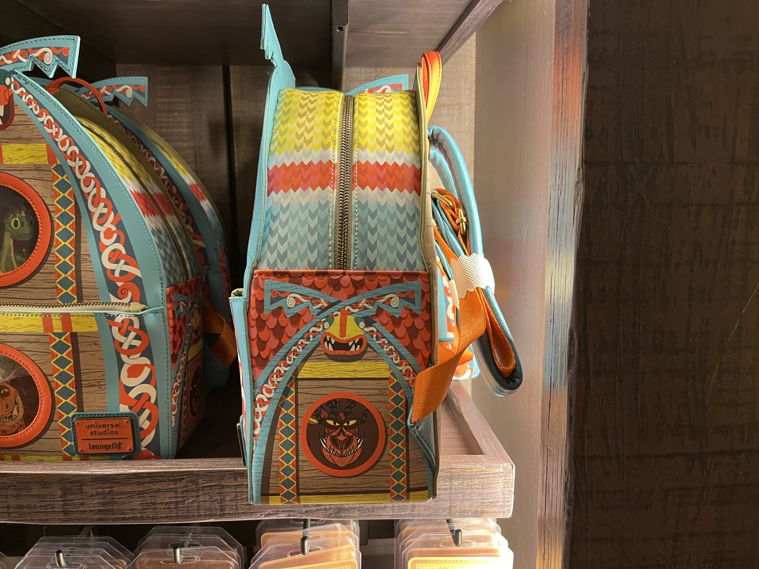 Colorful backpack with bold patterns and grinning cat face, displayed at Hiccup's Work Shop on a wooden shelf.