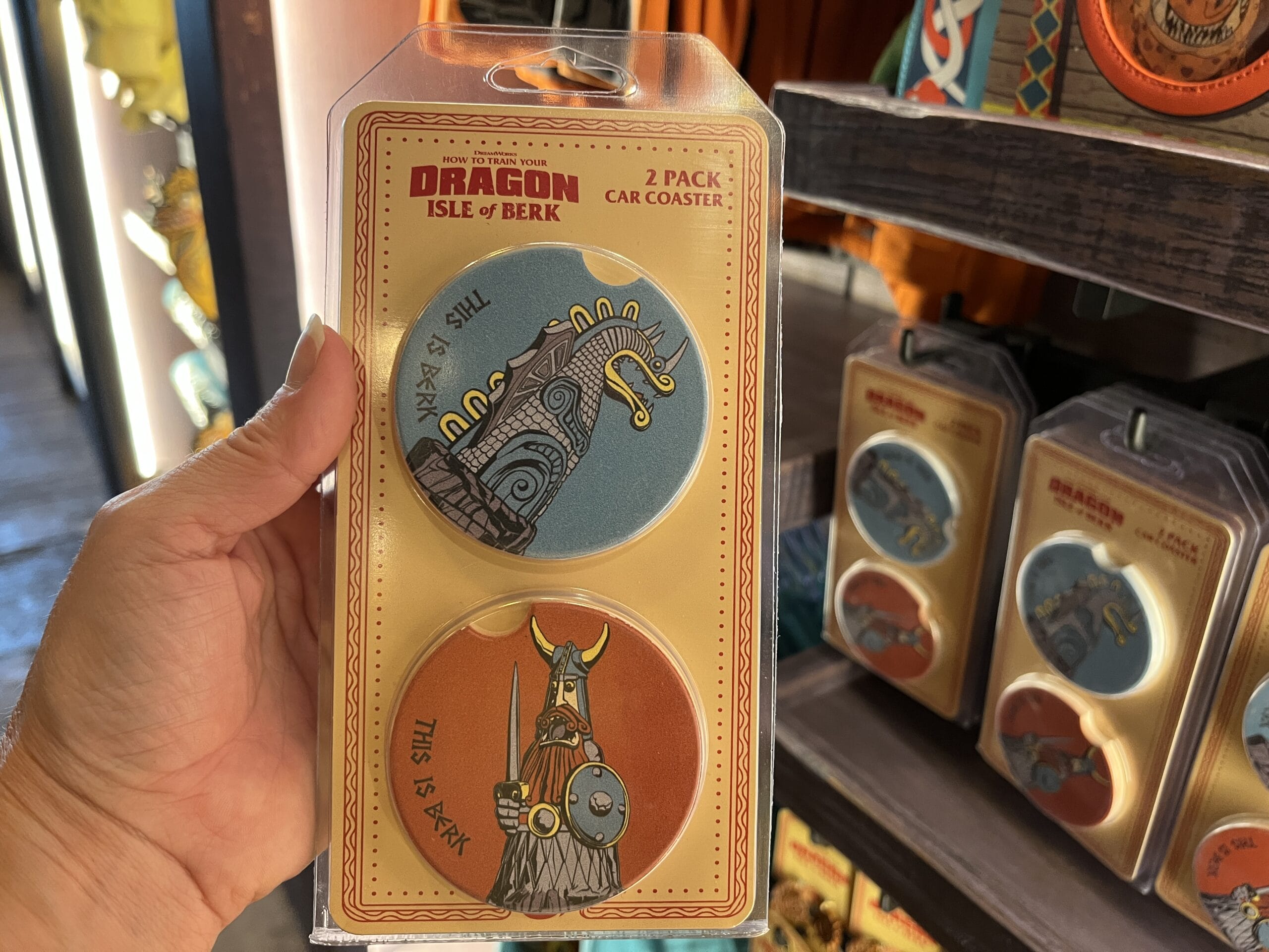 Hand holding a pack of two "Dragon Isle of Berk" car coasters from Hiccup's Work Shop with Viking and tower illustrations.