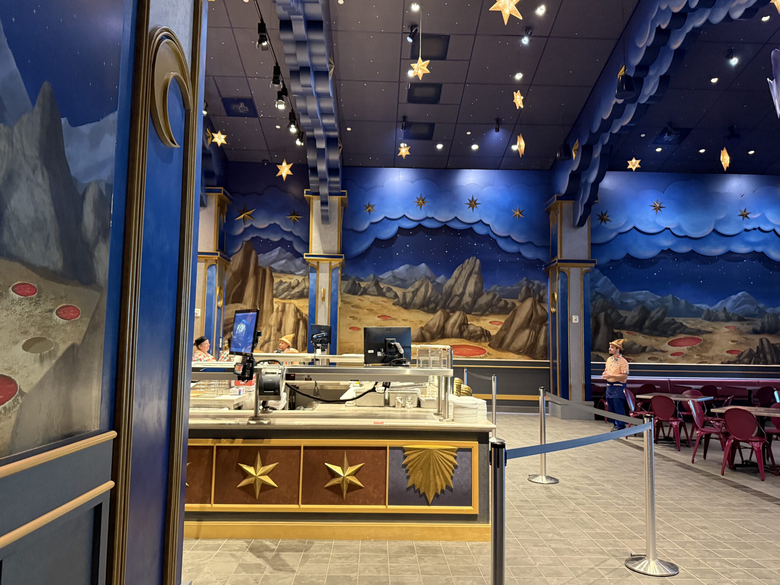 A space-themed Pizza Moon restaurant with star decor, blue walls, and a counter; a few people are inside.