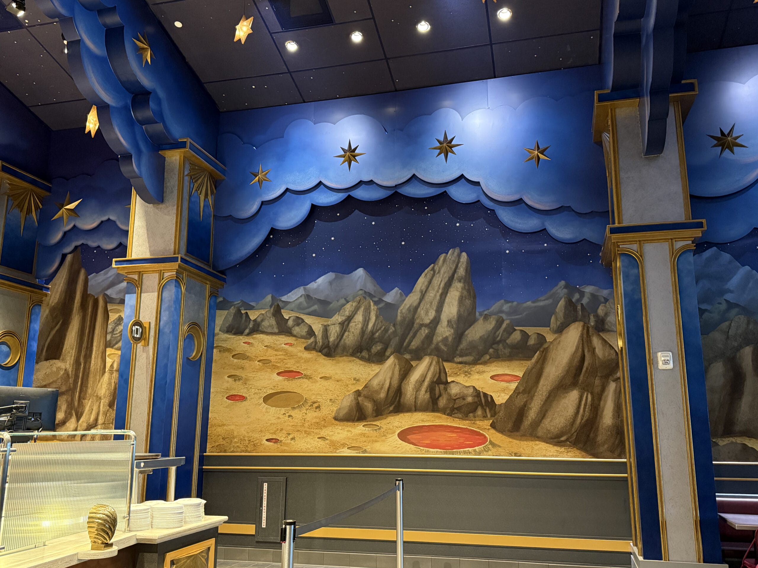 A restaurant interior at Pizza Moon Restaurant features a starry sky mural, rocky landscape, and red craters among blue columns.