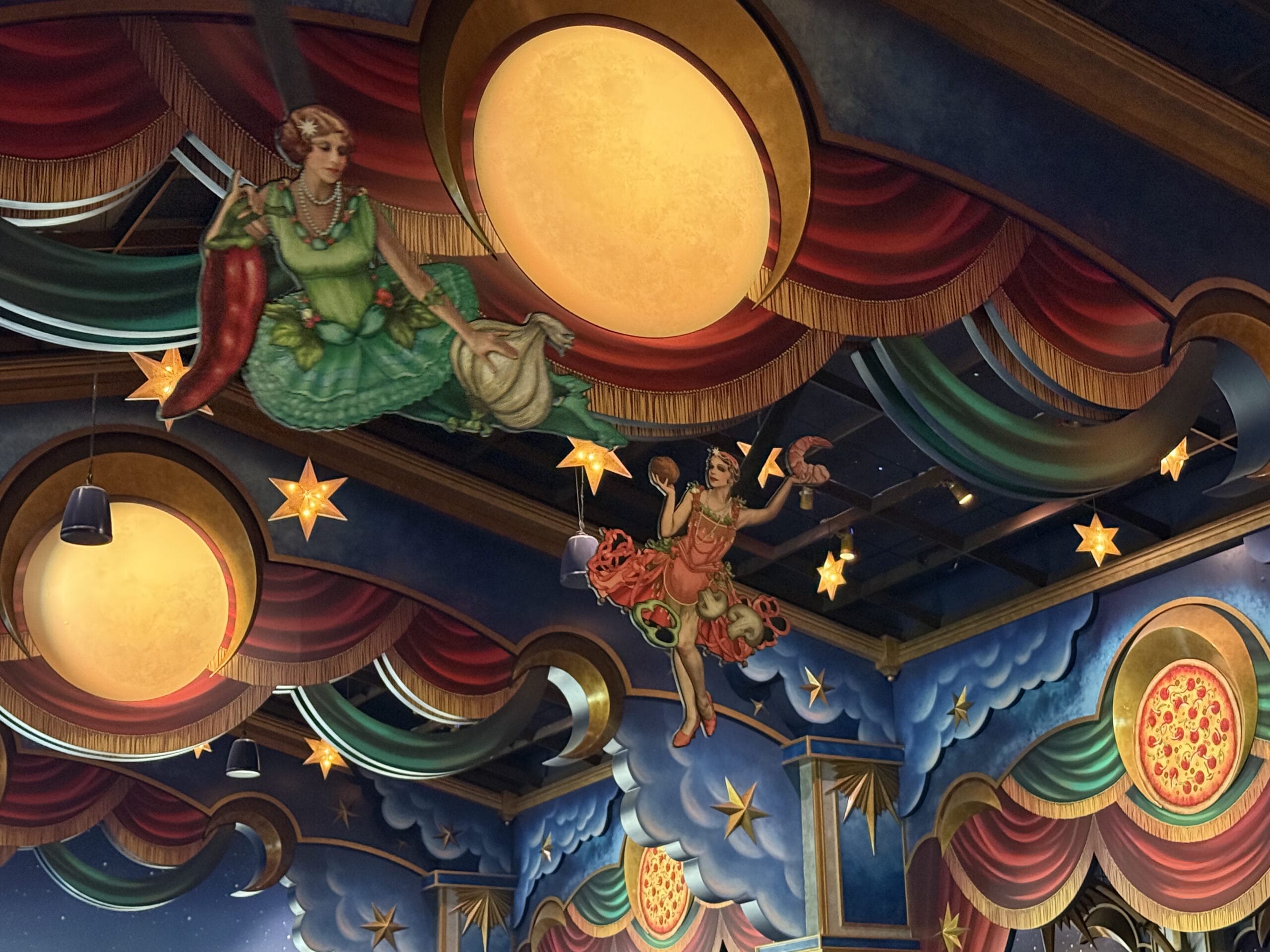 Whimsical ceiling art at Pizza Moon Restaurant features women with food, glowing lights, and painted pizzas in a celestial park sky.