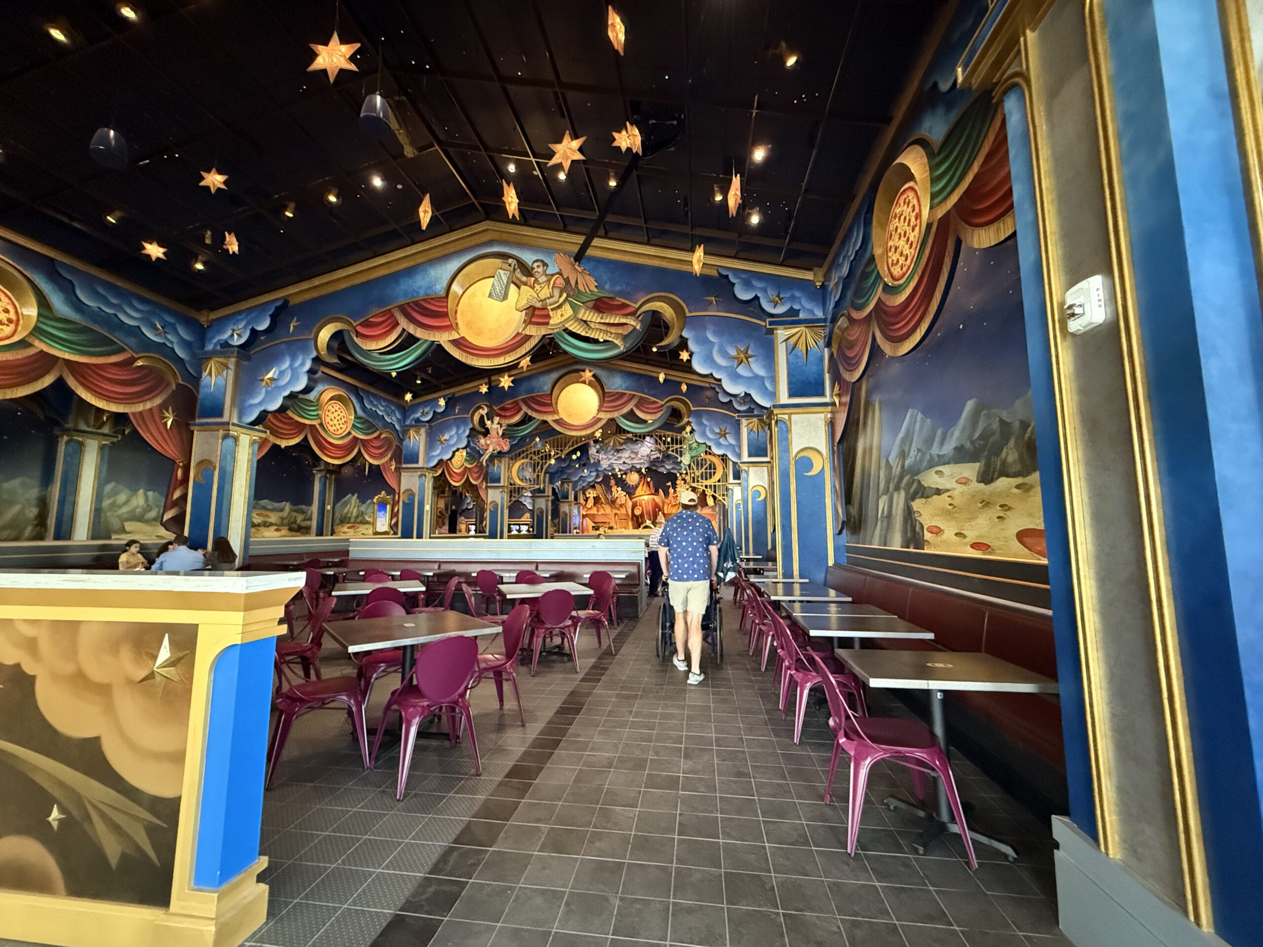 Colorful, space-themed Pizza Moon Restaurant interior with purple chairs, empty tables, and people walking toward a mural.