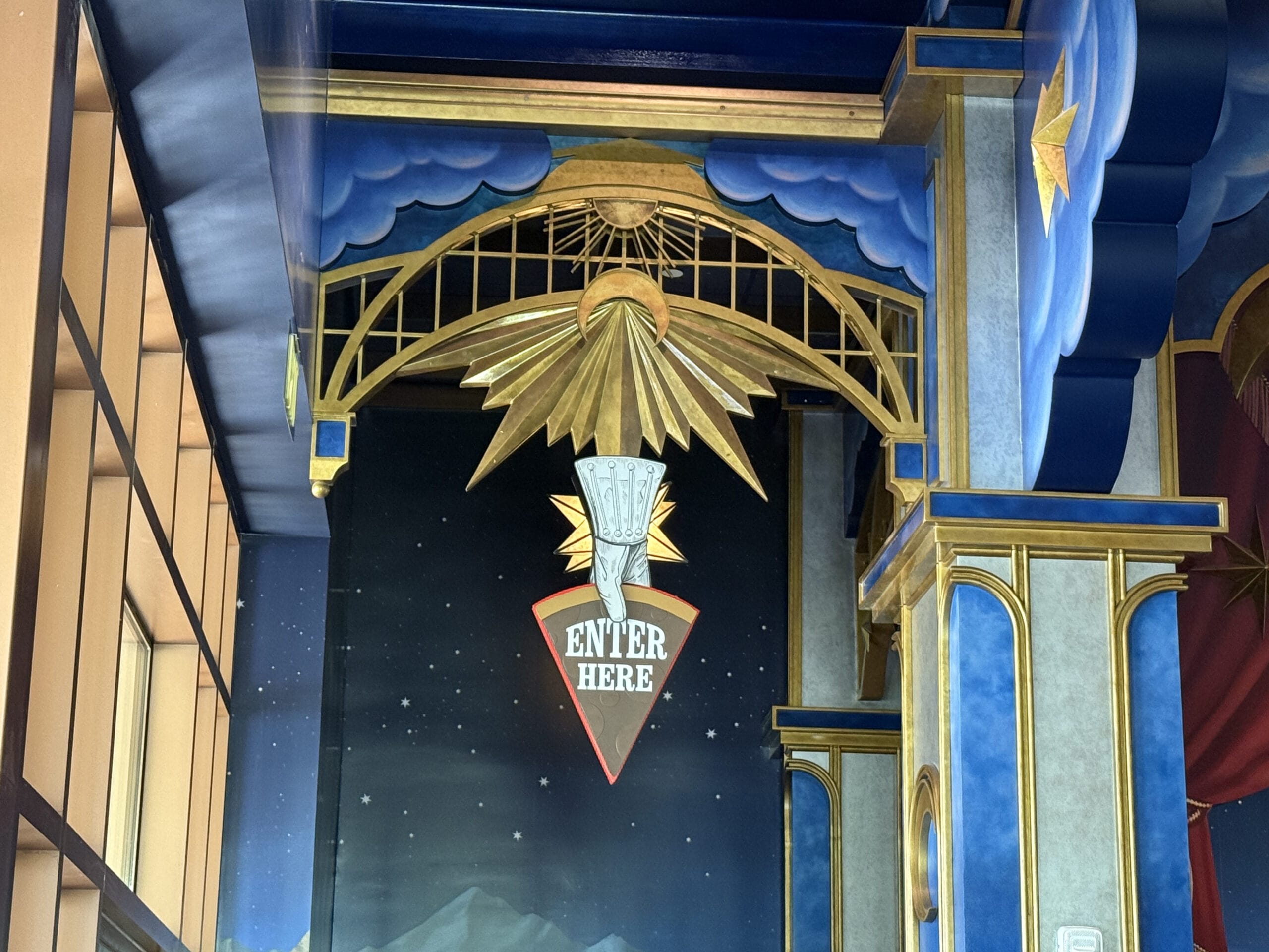 Gold art deco "Enter Here" sign with a pointing hand glows at Epic Universe’s Pizza Moon Restaurant in Celestial Park.