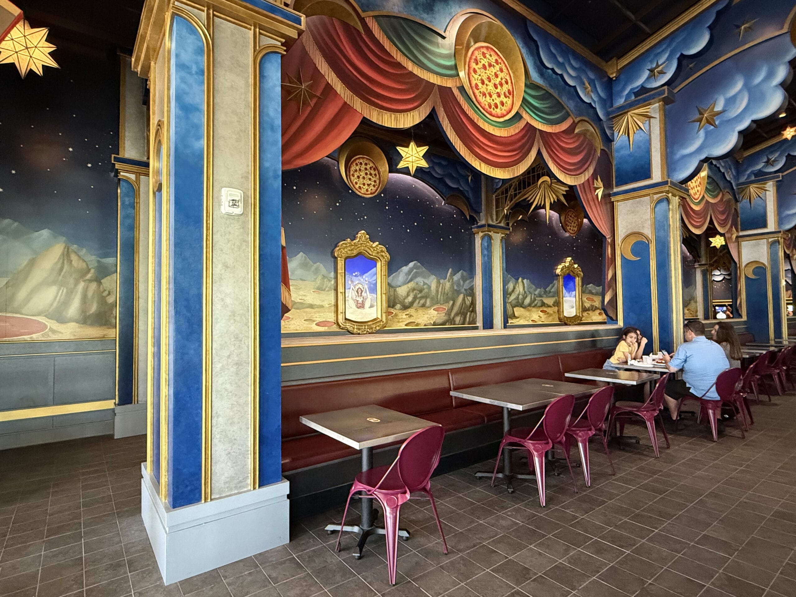 Colorful, whimsical Pizza Moon Restaurant with mountain murals; three people sit at a booth under celestial arches.