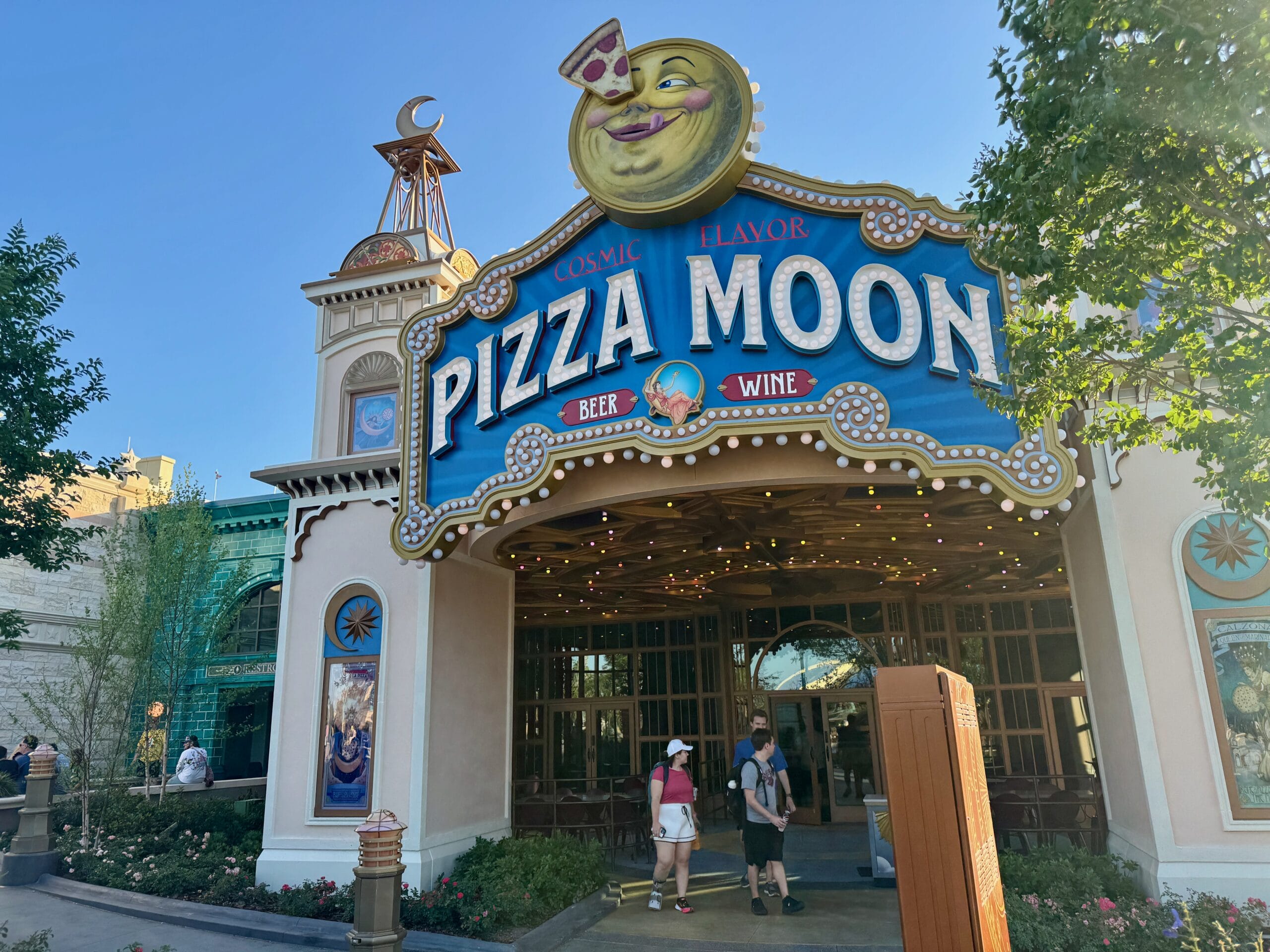 Colorful Pizza Moon Restaurant entrance near Celestial Park at Epic Universe, with a smiling moon sign and people passing by.
