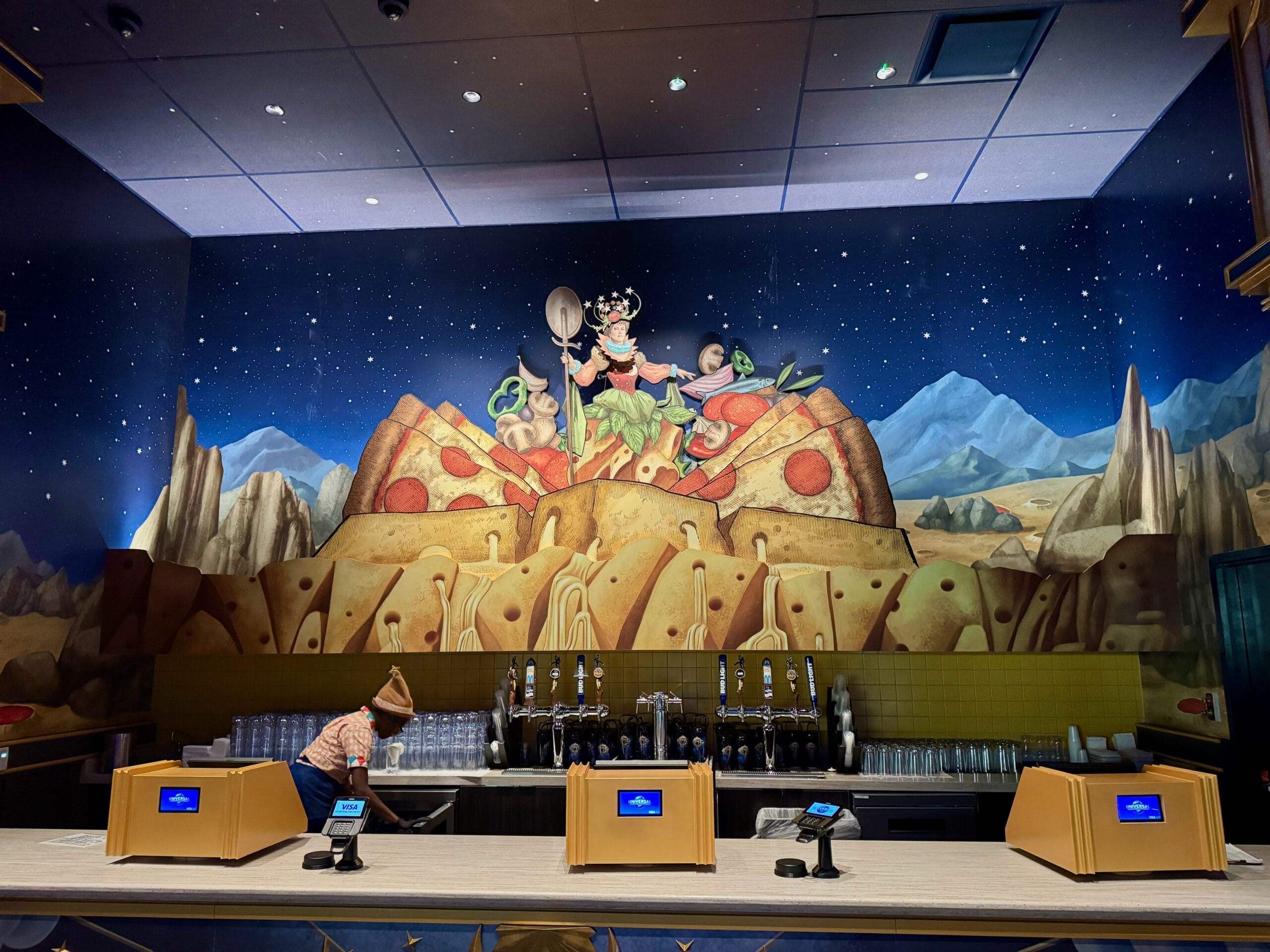 A mural of cartoon mice on a giant pizza decorates Pizza Moon Restaurant in Epic Universe’s Celestial Park; staff sets up counters.