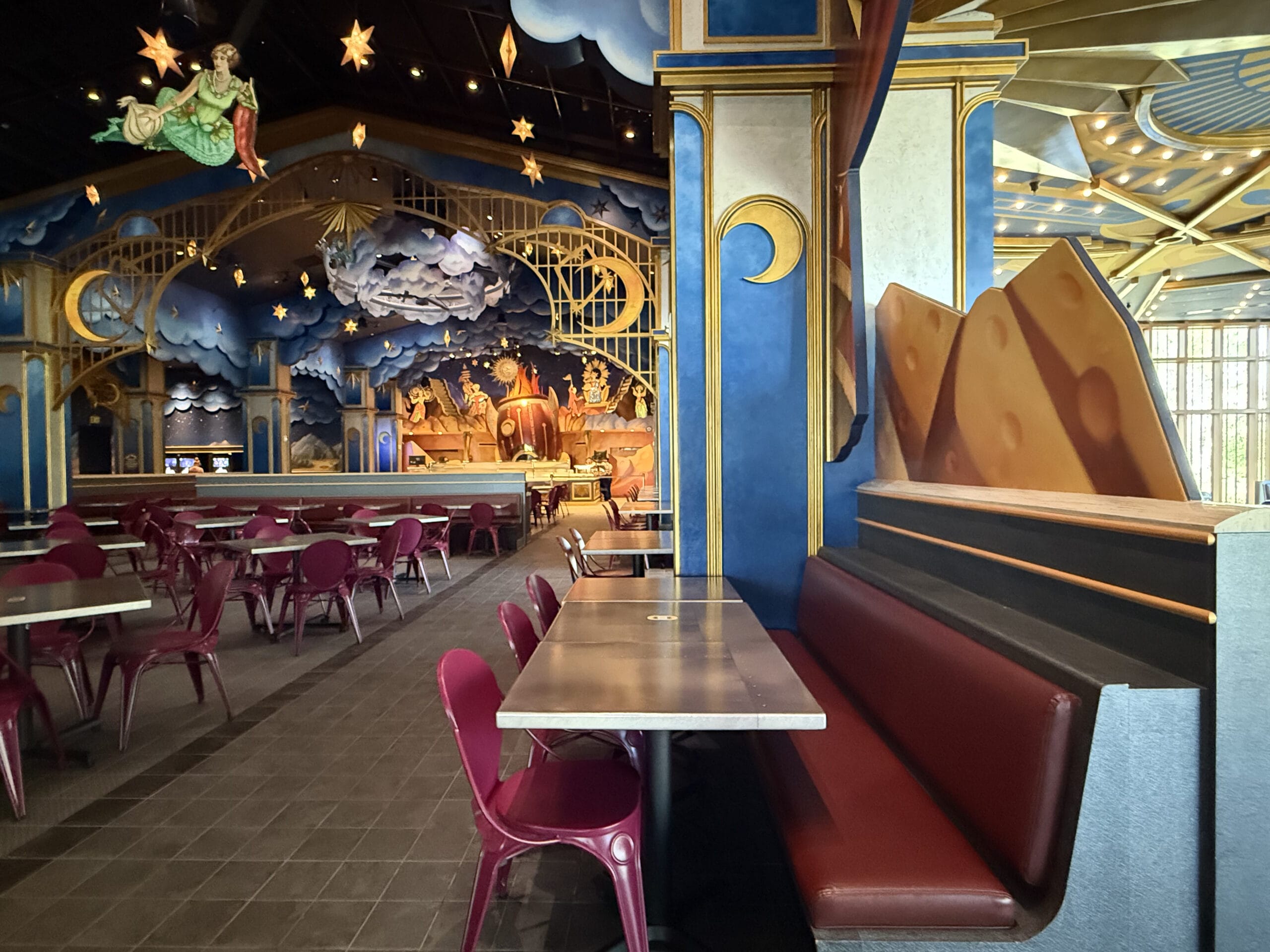 A whimsical, star-themed Pizza Moon Restaurant with red chairs, booths, and a fantasy mural of clouds and theatrical figures.