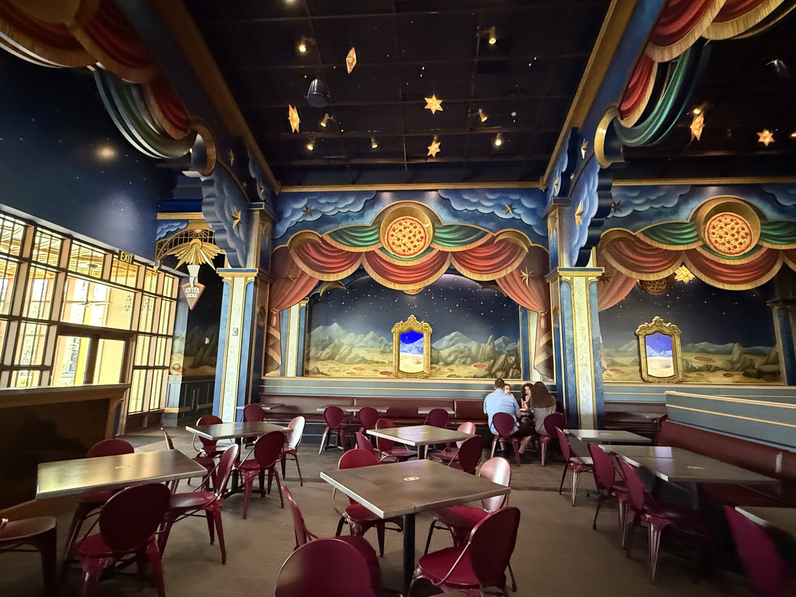 Colorful, theatrical café at Epic Universe with stage-like murals, red chairs, and two people seated in the background.