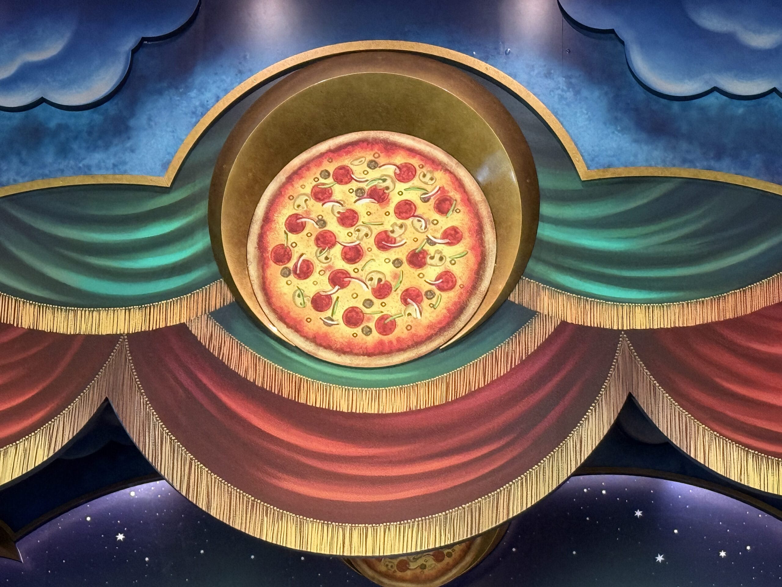 A painted pizza shines on a colorful, theatrical backdrop at Pizza Moon Restaurant in Epic Universe’s Celestial Park.