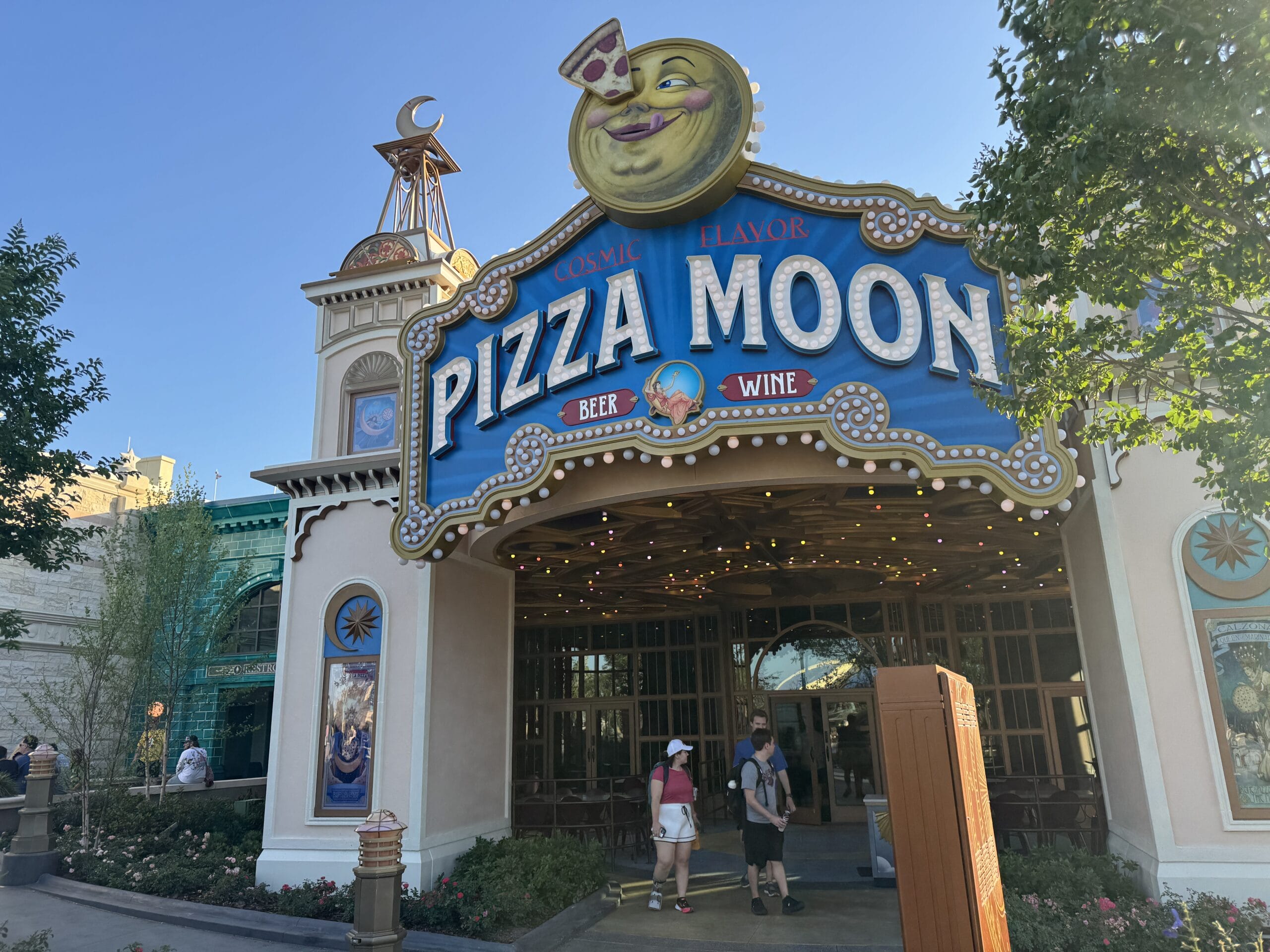 Entrance to Pizza Moon Restaurant at Epic Universe, with a smiling moon sign, near Celestial Park and people strolling by.
