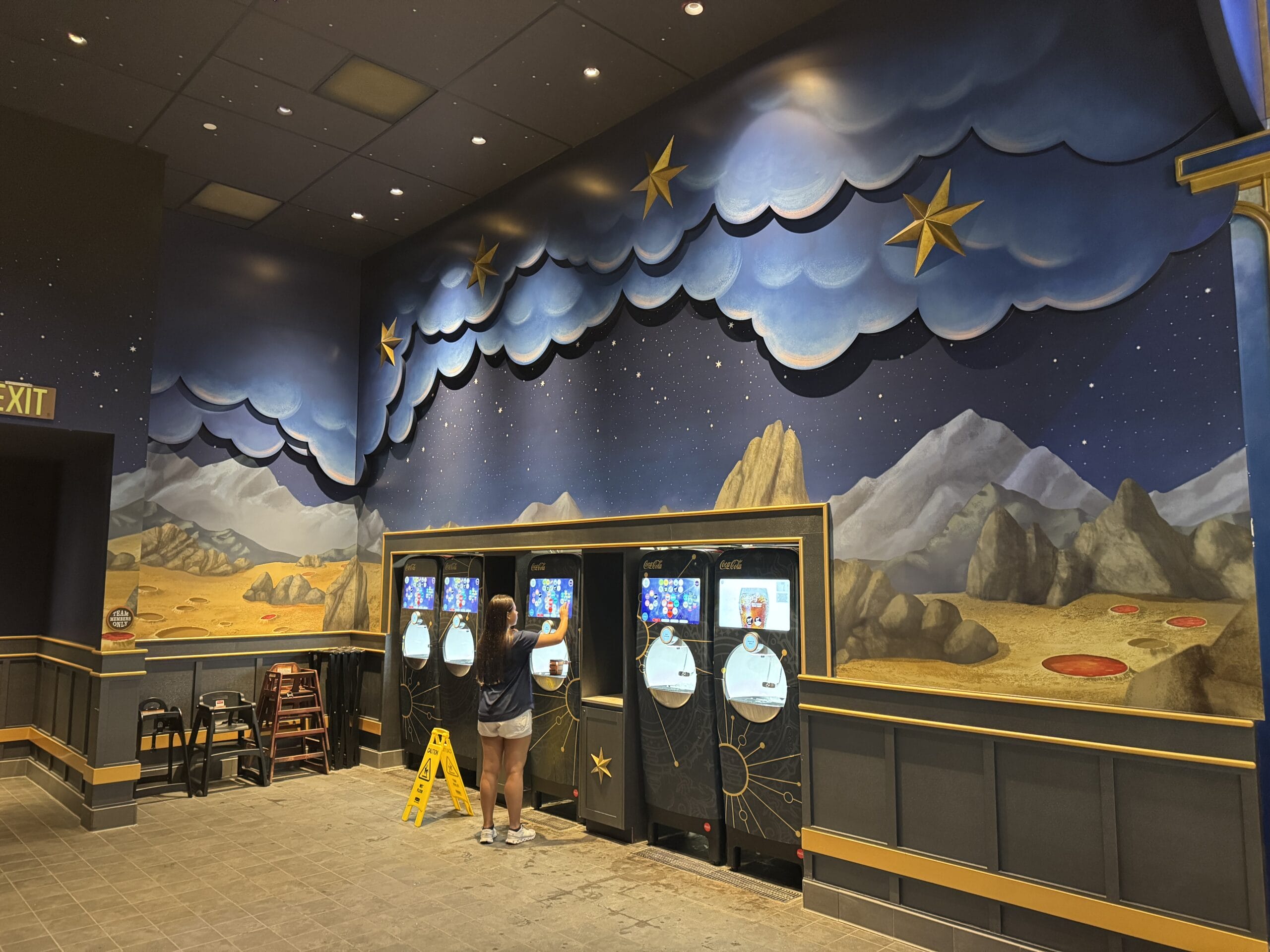 A person uses a self-service kiosk beneath a mural in the Epic Universe Celestial Park indoor space.