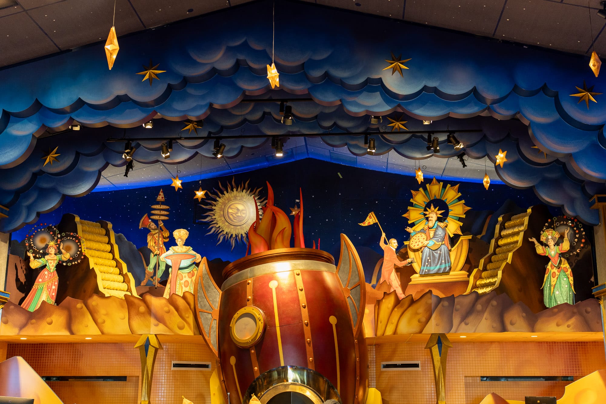 A whimsical stage set in Celestial Park with sun motifs, colorful figures, and a rocket ship beside Pizza Moon.