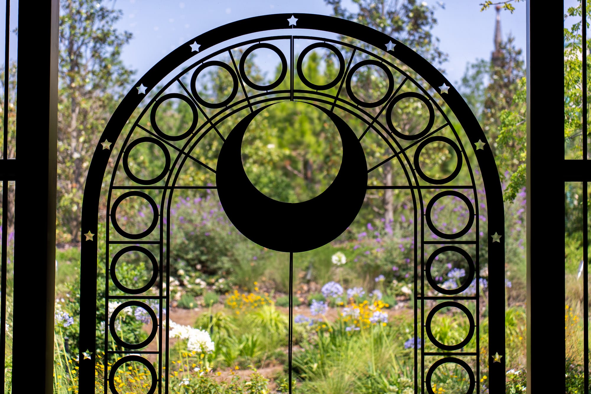 Black iron gate with crescent moon and circles, overlooking Celestial Park’s lush garden of flowers and trees.