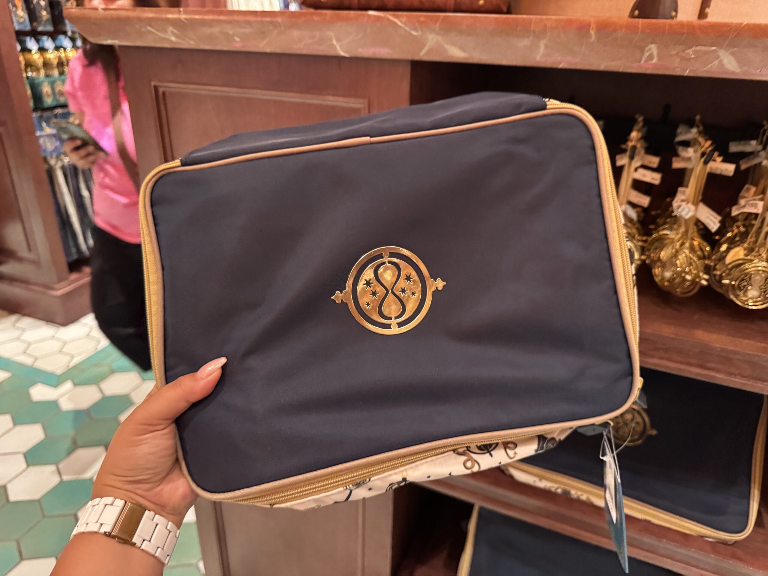 A hand holds a navy blue zippered bag with a gold emblem, perfect for Wizarding World merchandise shopping in-store.