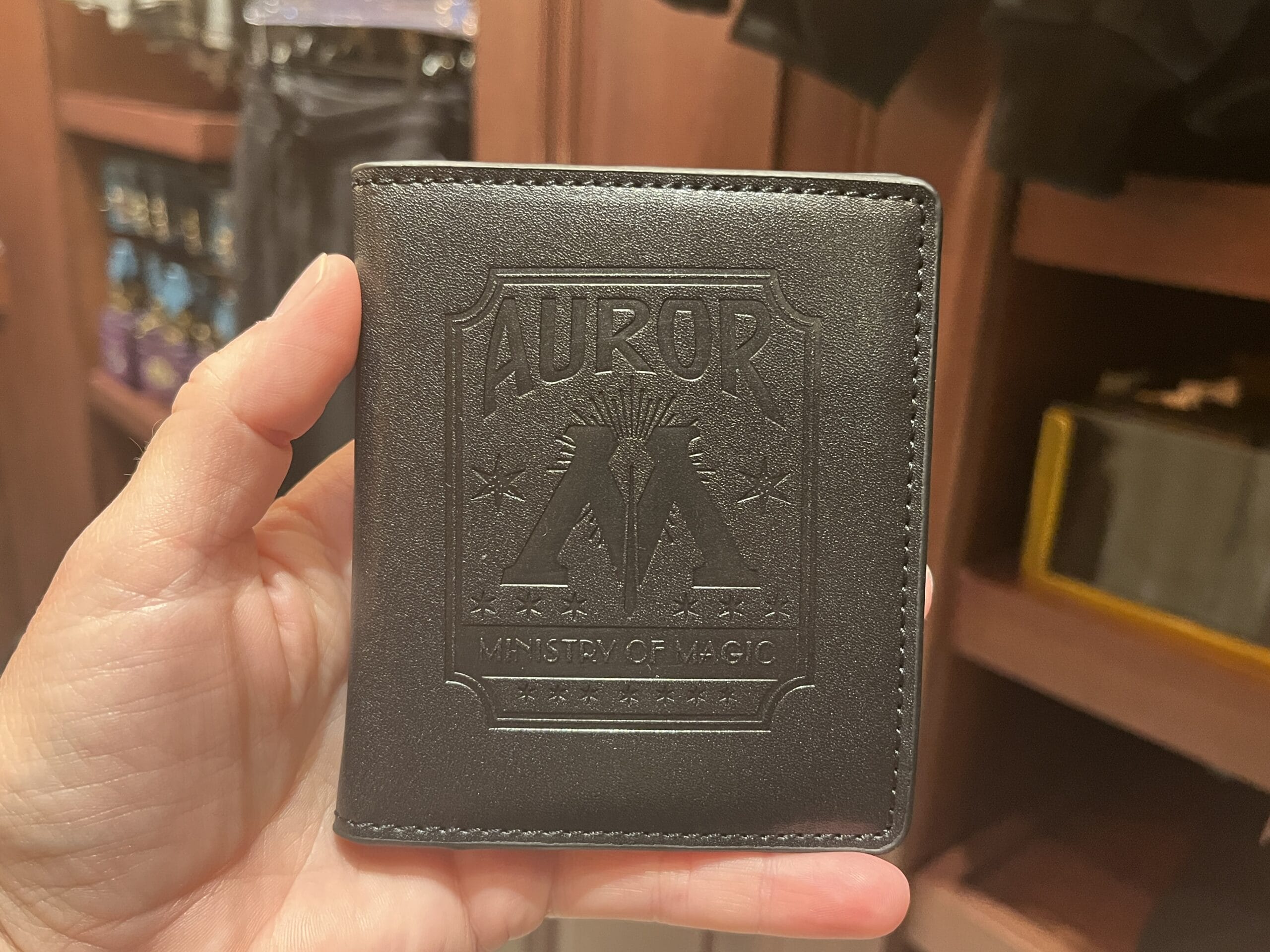 A hand holds Wizarding World of Harry Potter merchandise—a black wallet embossed "Auror Ministry of Magic" and a stylized "M.