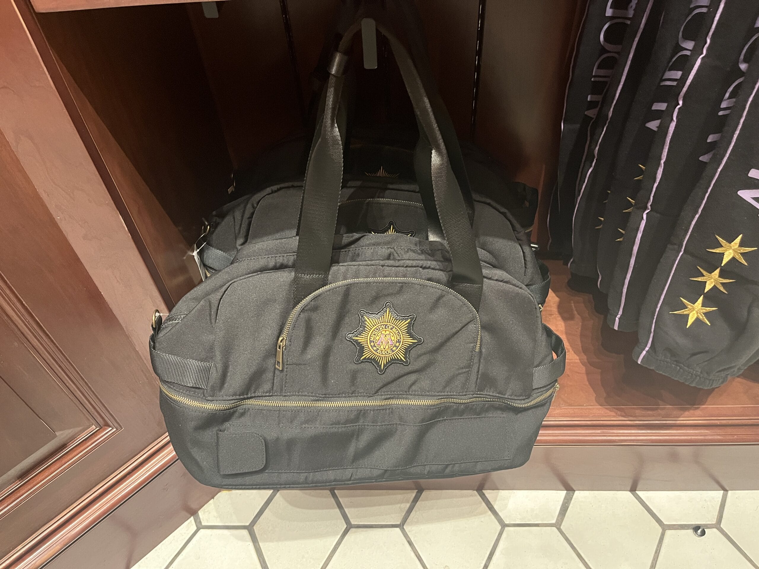 Black duffel bag with gold embroidered star, displayed on a shelf next to Harry Potter merchandise and black clothing.