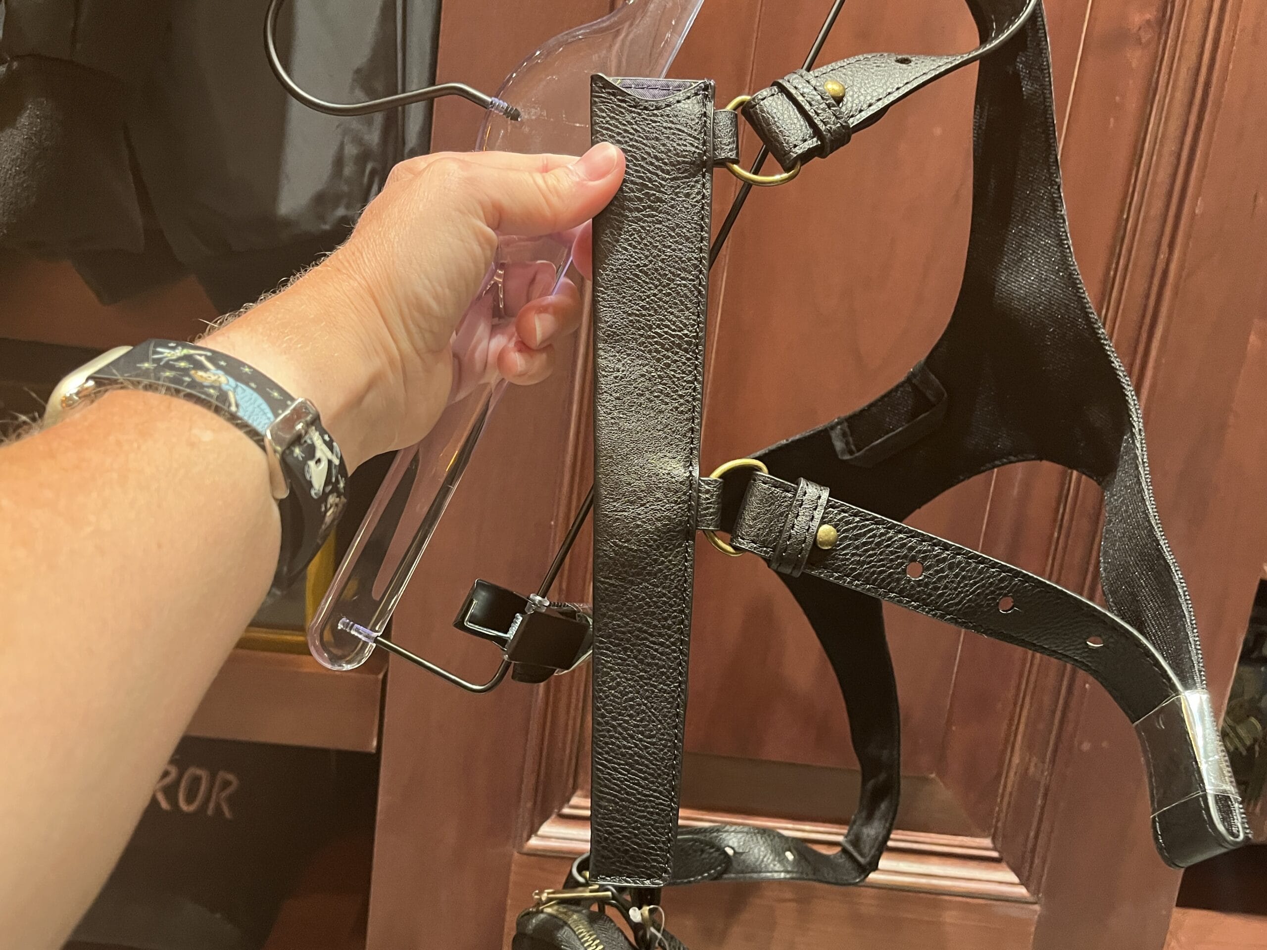 A hand holds a black leather harness—Epic Universe’s latest Wizarding World of Harry Potter merchandise—in front of wood.