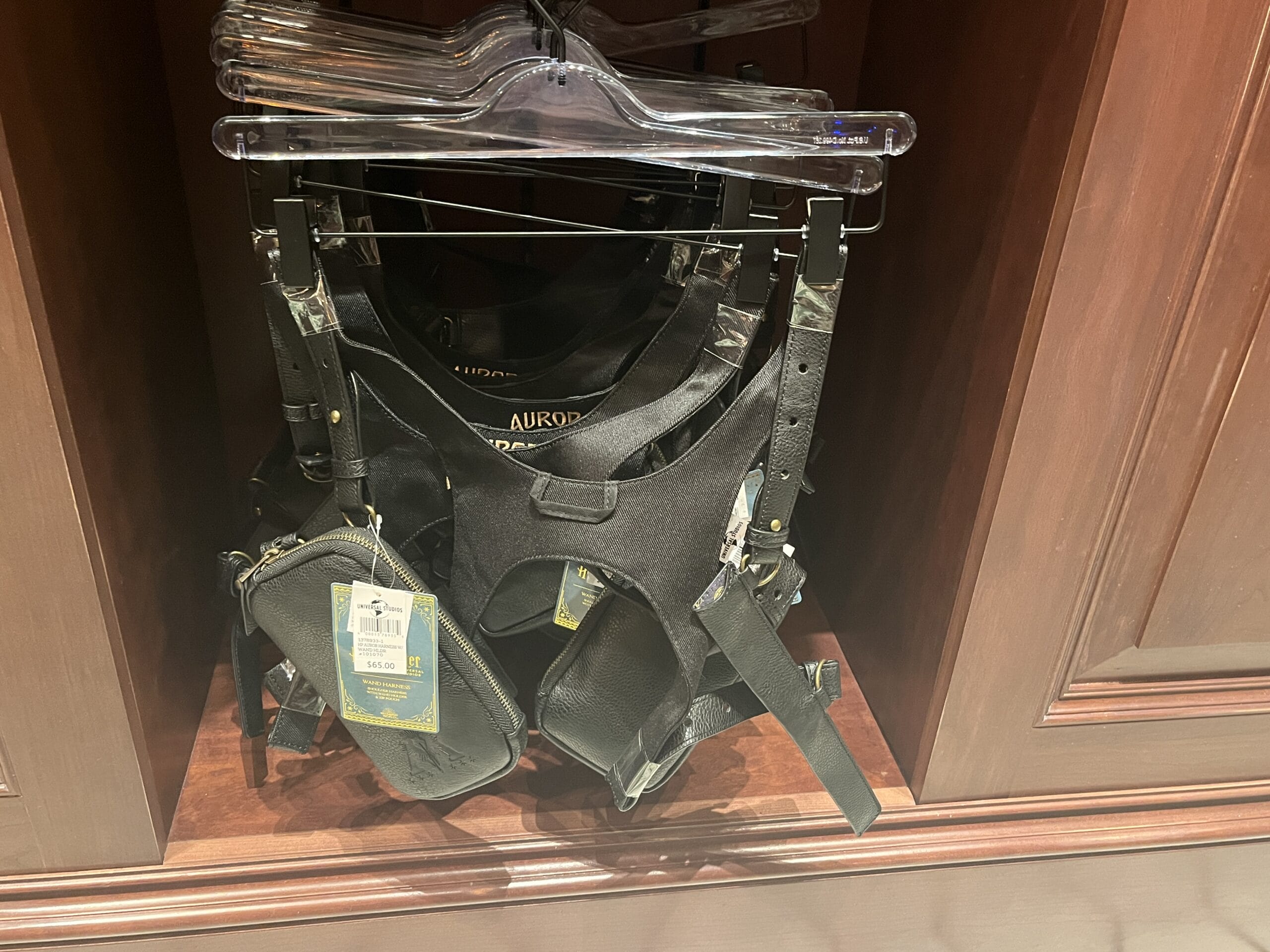 Black leather harnesses with merchandise prices hang on clear plastic hangers in a wooden display case.