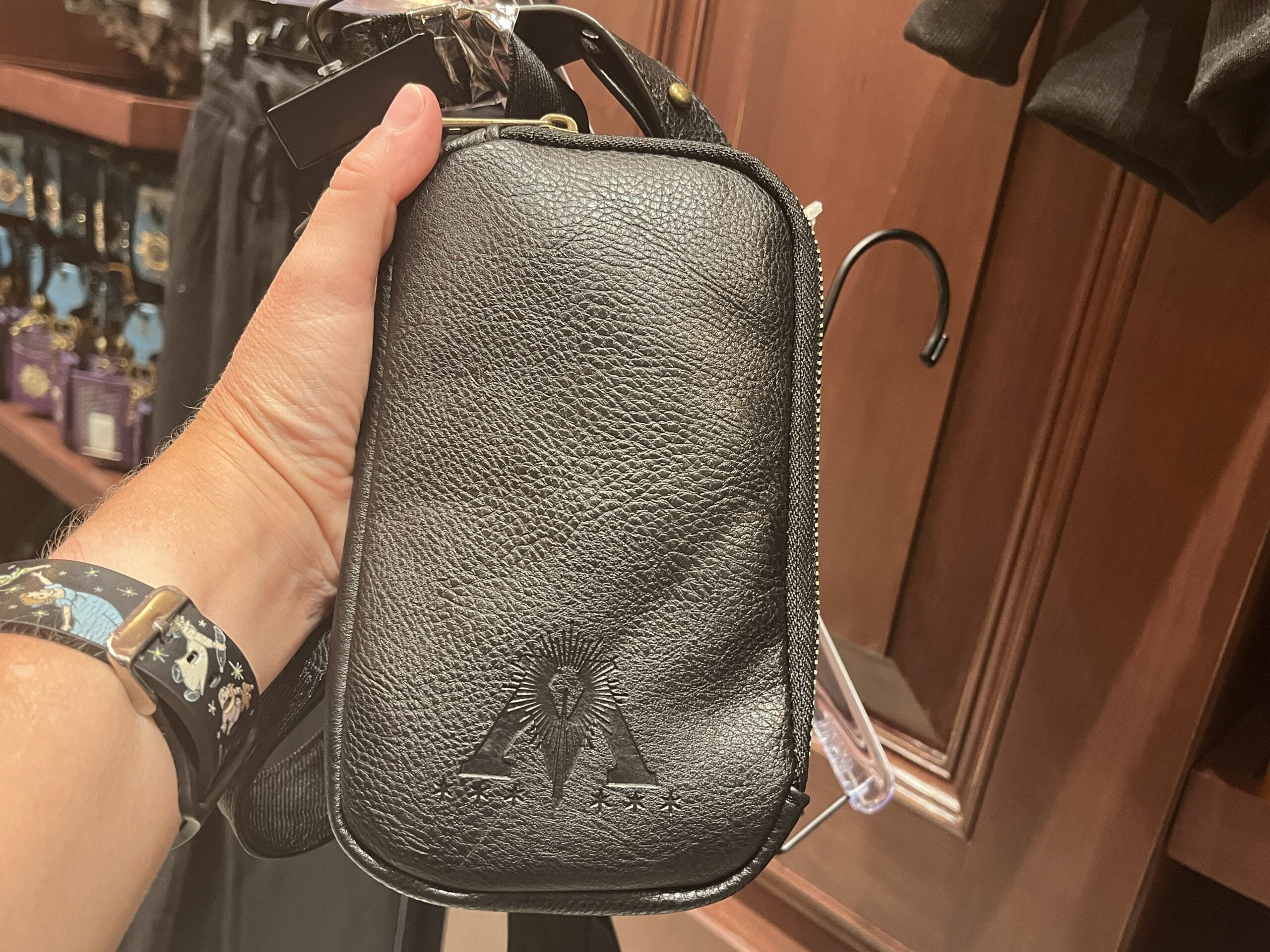 A hand holds a black leather pouch with a subtle embossed design—unique Tour En Floo merchandise in the store's wooden shelves.