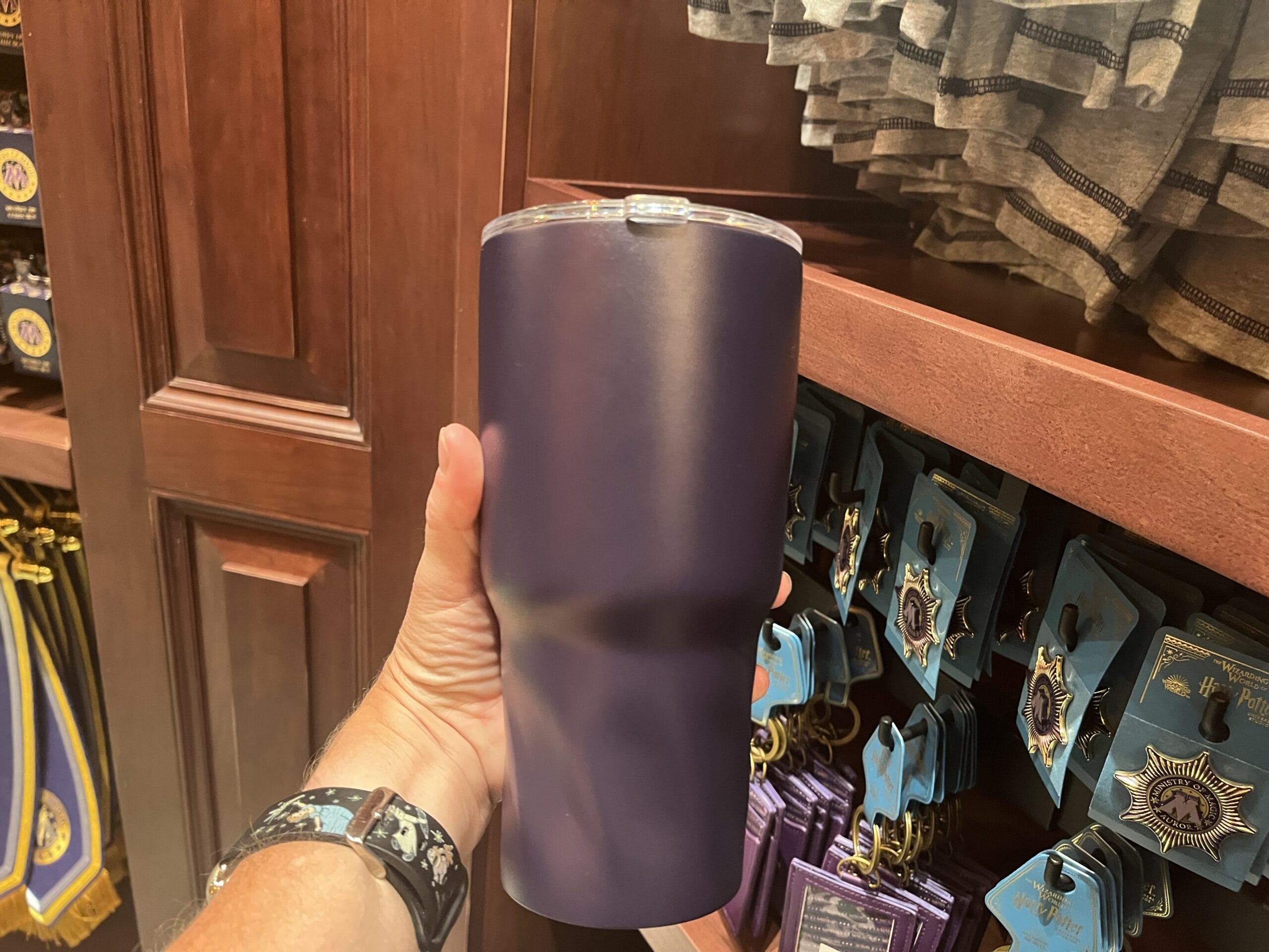 A hand holds a tall, matte purple tumbler in a store at Epic Universe, with pins and cloth near Tour En Floo merchandise.