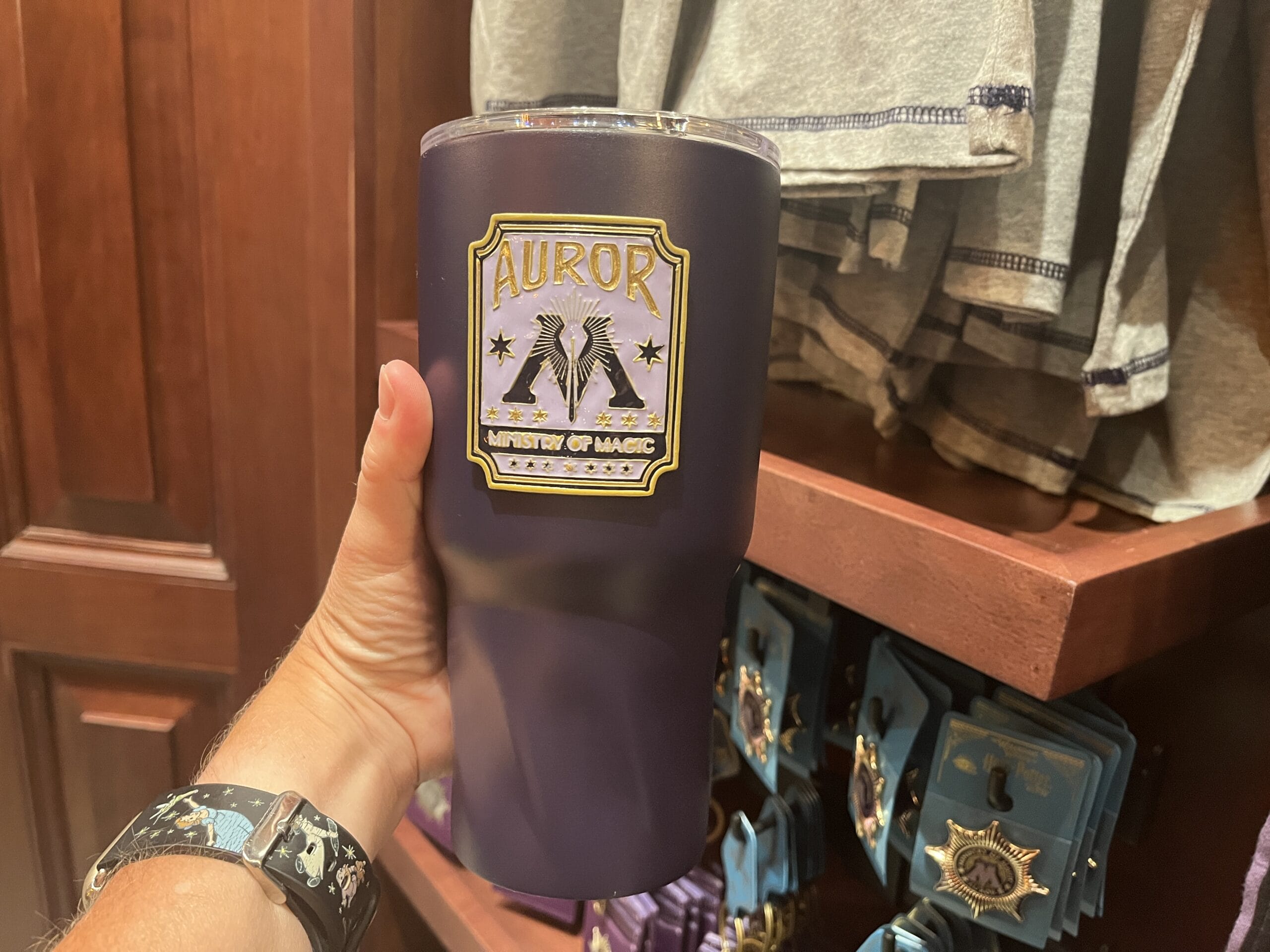 A hand holds a dark tumbler labeled "Auror Ministry of Magic" amid Wizarding World of Harry Potter merchandise in a store.