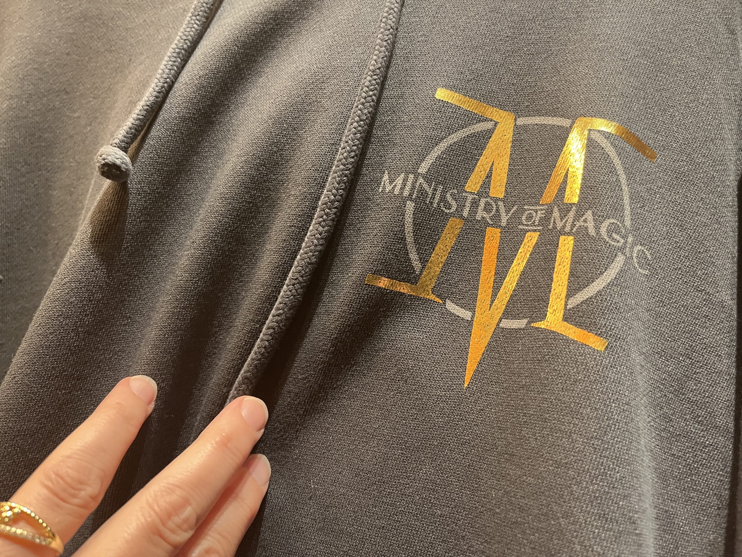 A hand touches a gray hoodie with a gold "Ministry of Magic" logo—perfect Wizarding World merchandise for fans.