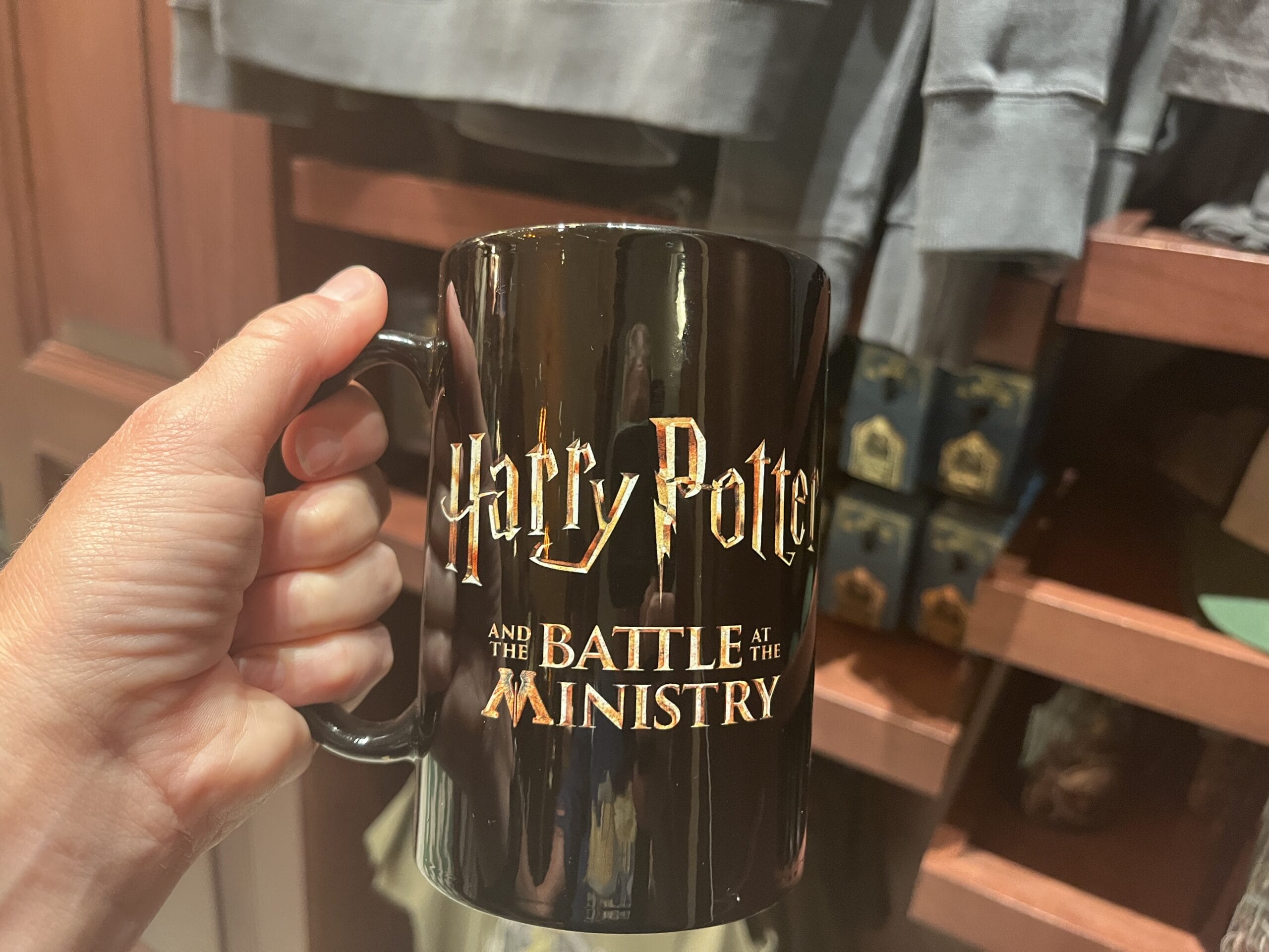 A hand holds a black mug with gold text—a magical piece of Wizarding World of Harry Potter merchandise.