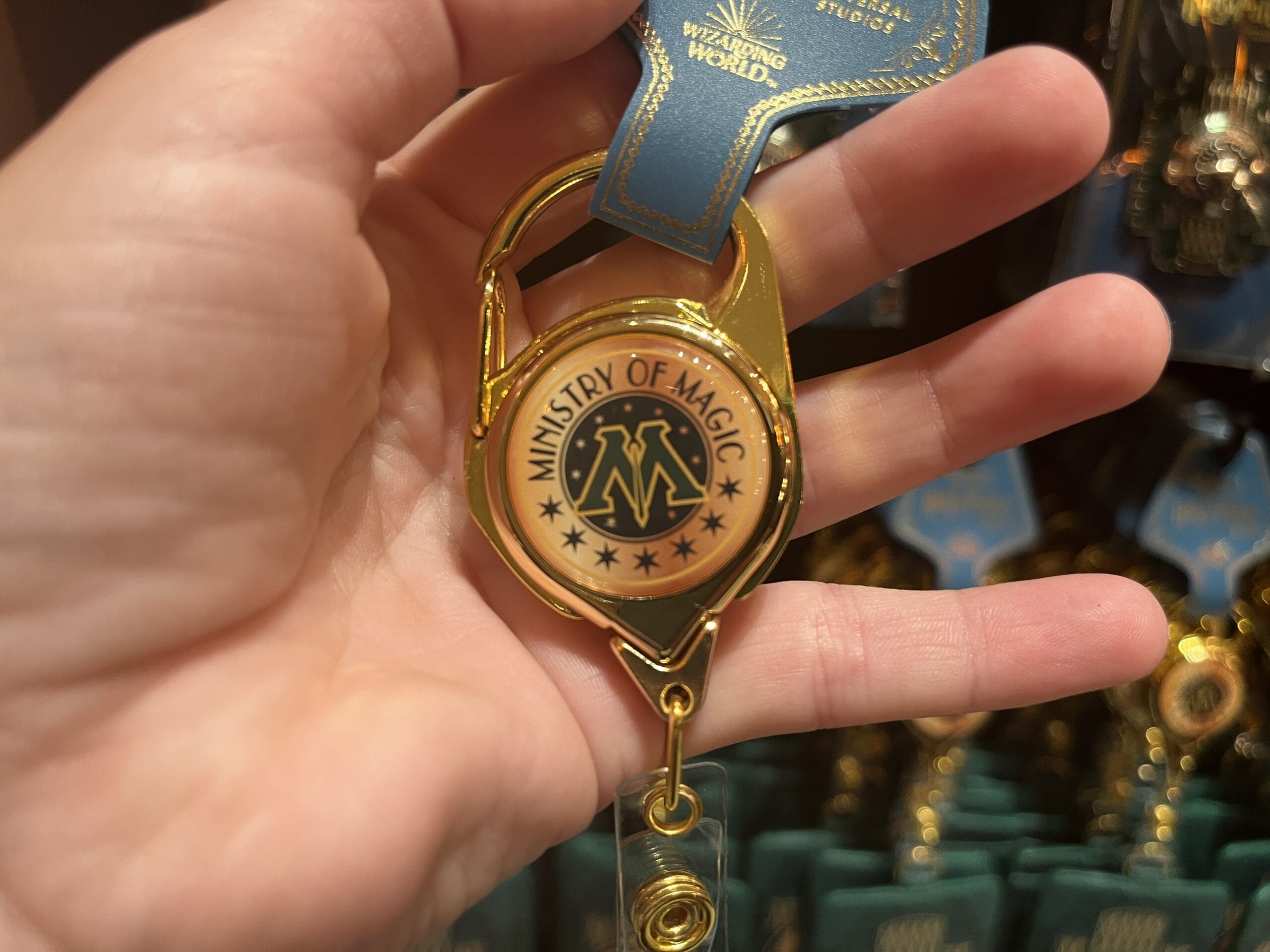 A hand holds a gold "Ministry of Magic" keychain, exclusive Wizarding World merchandise from the Harry Potter tour.