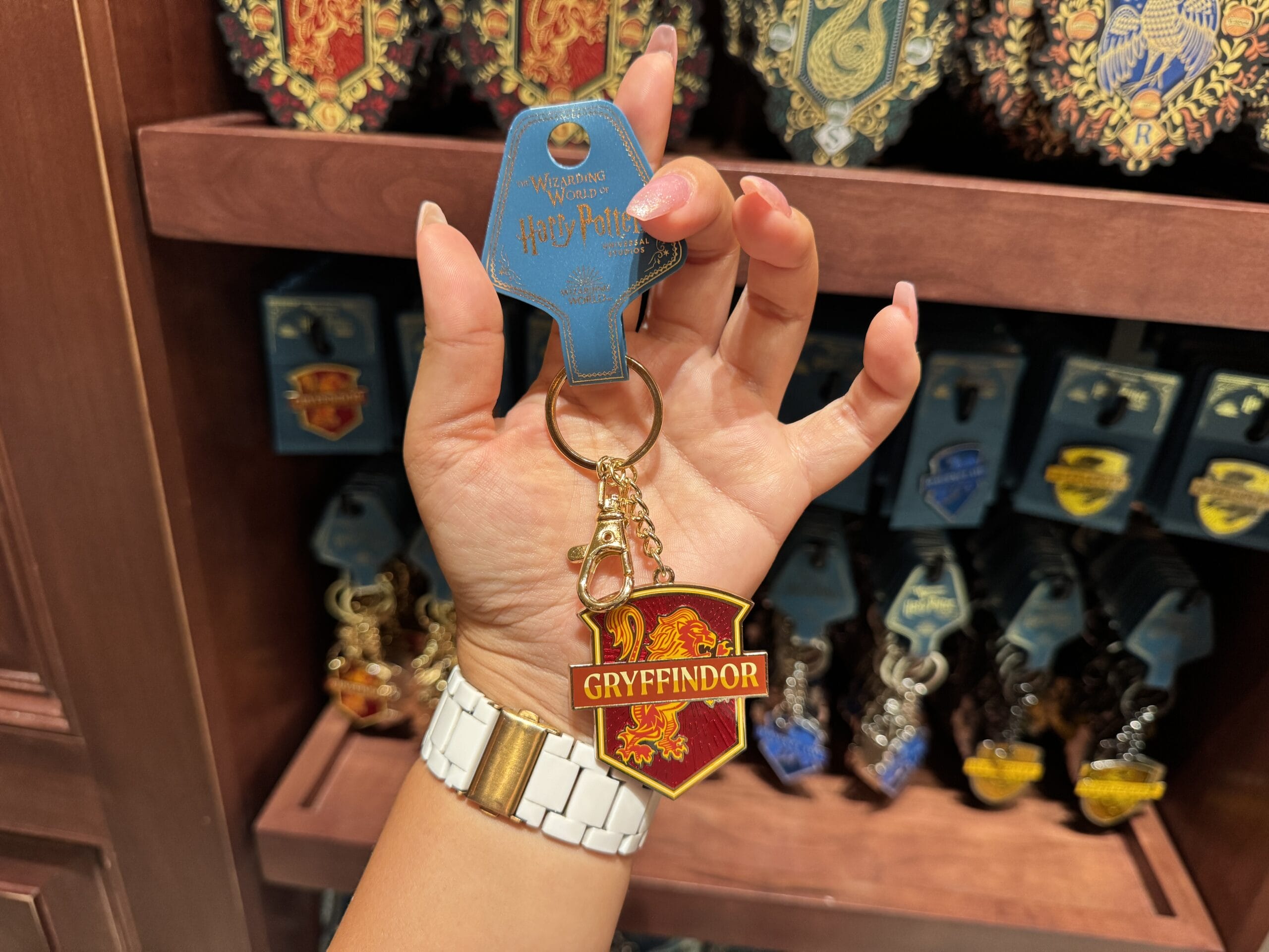 A hand holds a Gryffindor keychain in a Wizarding World shop, with Harry Potter merchandise from other houses in the background.