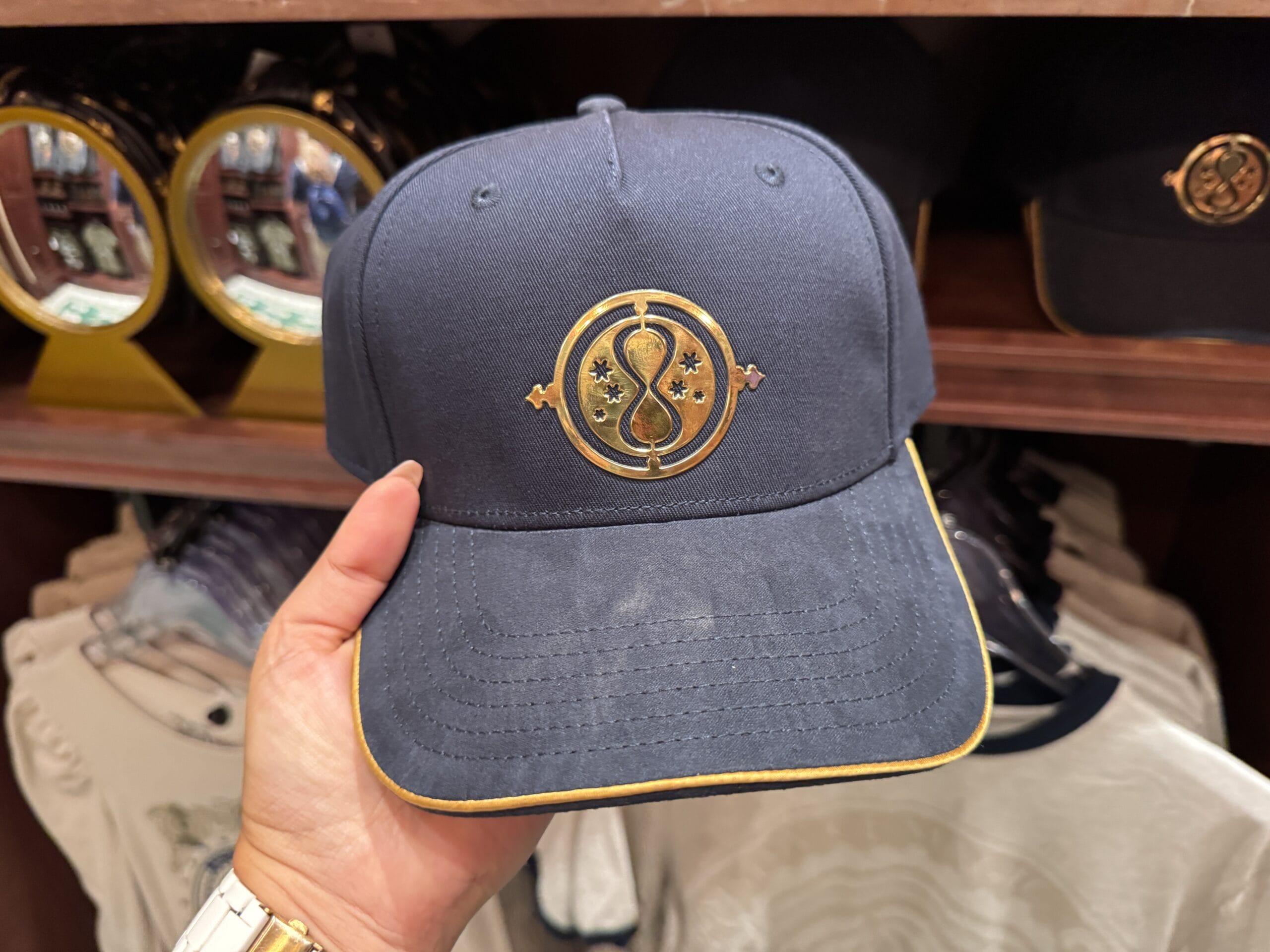 A hand holds a navy blue hat with a gold emblem, reminiscent of Wizarding World style, displayed in a retail store.