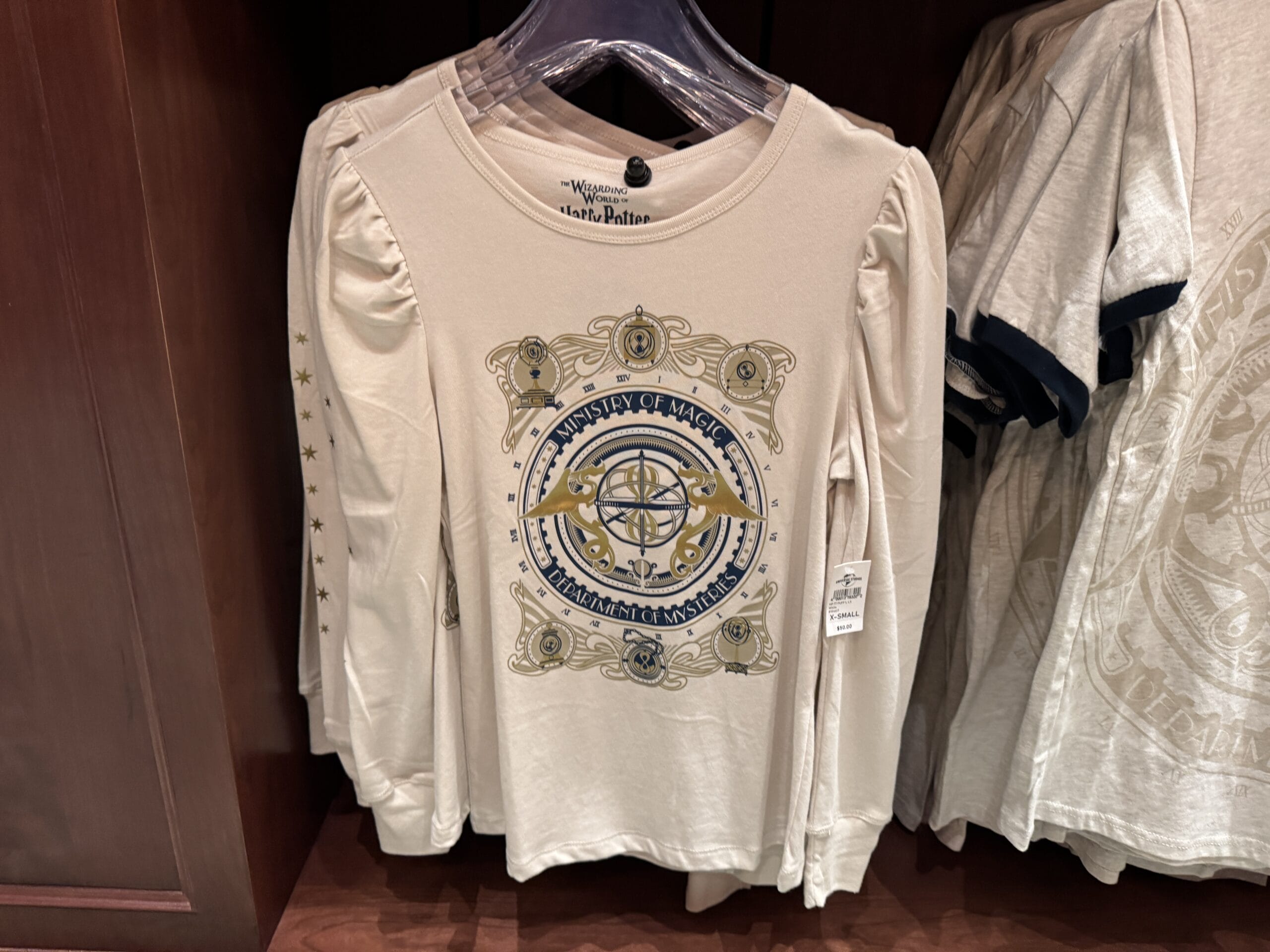 White long-sleeve "Ministry of Magic" shirt, a Wizarding World of Harry Potter merchandise item, hangs on a store rack.