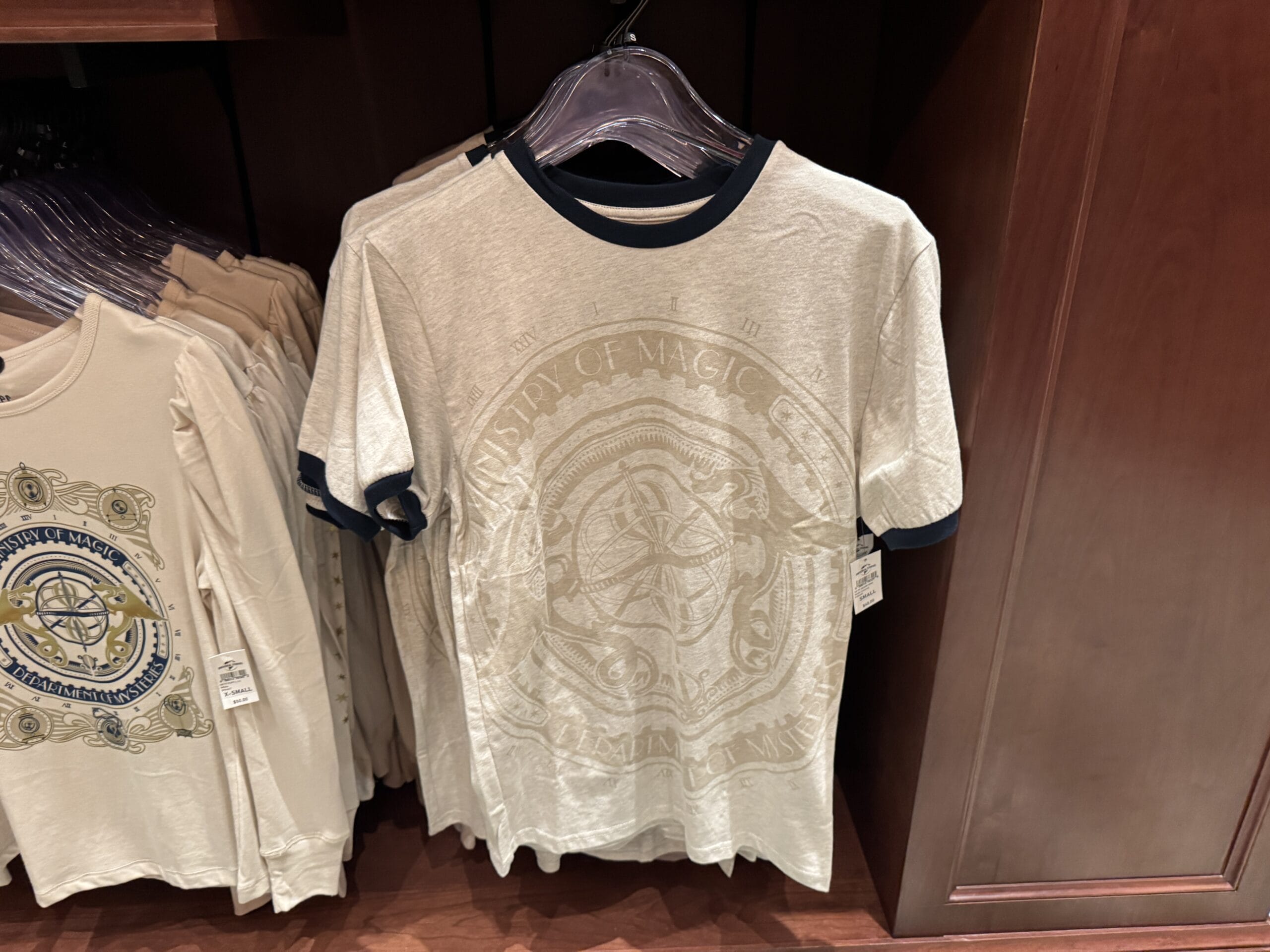 Beige T-shirt with dark trim, a faded magical emblem—perfect Wizarding World of Harry Potter merchandise—hanging on a rack.