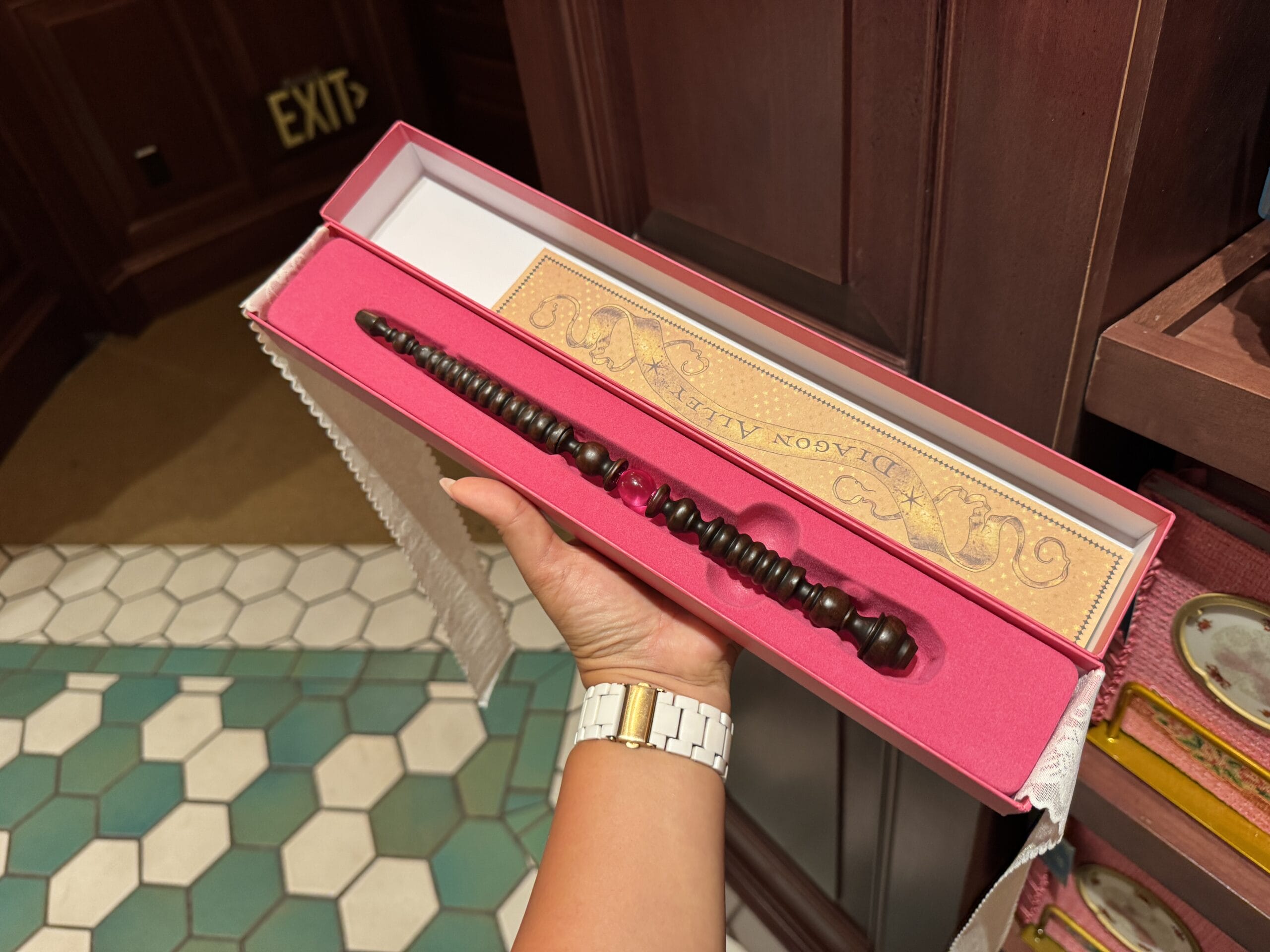 A hand holds a dark wooden wand, Wizarding World of Harry Potter merchandise, in a pink Diagon Alley box at a store.